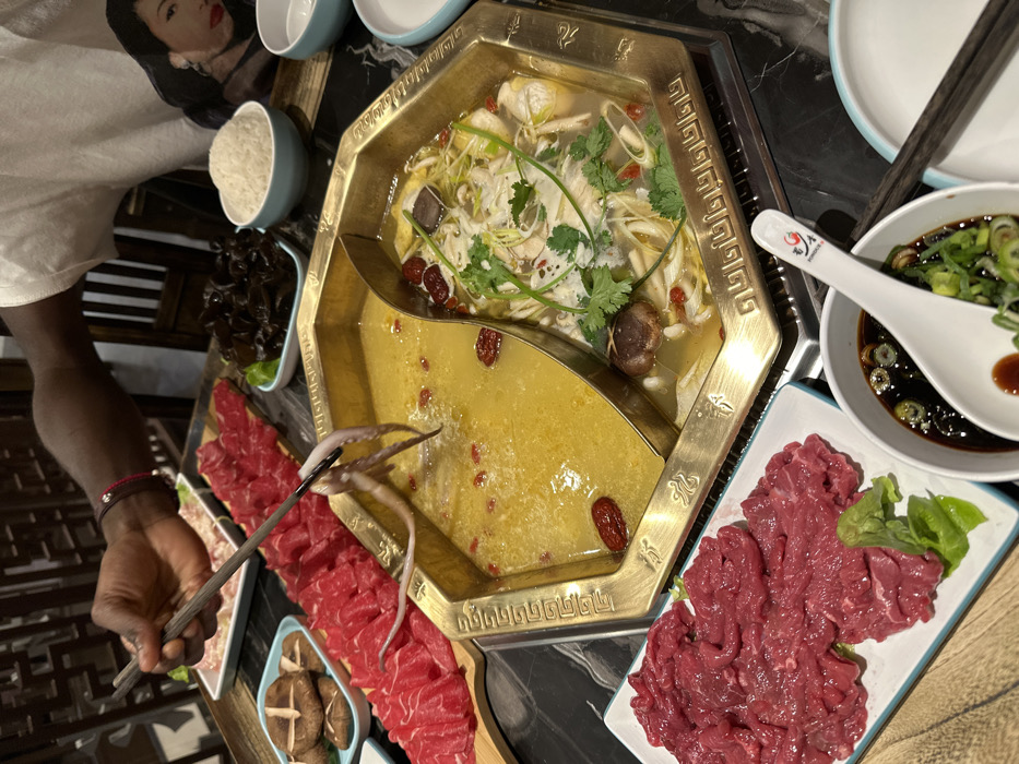 The scene depicts a close-up, overhead view of a hot pot dining experience within a restaurant in Paris, France. A dark-skinned arm and hand, belonging to an individual wearing a white t-shirt featuring a graphic of a woman's face, uses dark chopsticks to immerse a whole, small octopus into the simmering yellowish broth of the left compartment of a brass-colored, divided hot pot. This broth contains visible red dates. The right compartment of the hot pot holds a lighter, clear broth containing sliced poultry or fish, shiitake mushrooms, leeks, cilantro, and red goji berries.
The dark, marble-patterned tabletop is laden with various raw ingredients and condiments. To the left, a long wooden board displays multiple portions of thinly sliced raw red meat. A white rectangular plate in the lower-left foreground also contains a pile of thinly sliced raw red meat, garnished with a few green leaves. Other plates hold whole shiitake mushrooms and rehydrated dark wood ear mushrooms. Several white and light blue ceramic bowls are visible, including bowls of white rice. In the lower right, a white bowl holds a dark dipping sauce, garnished with chopped green onions, containing a white ceramic spoon with a red logo. The background features dark wooden latticework, suggesting traditional restaurant decor.
