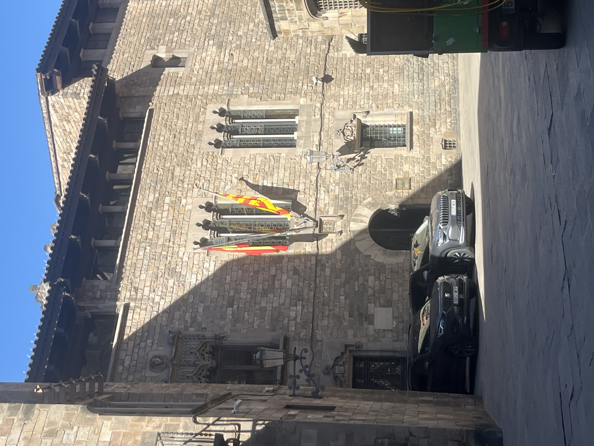 The image shows a building facade in Barcelona, Spain. The building is constructed of tan stone blocks. A Catalan flag hangs from a bracket on the wall between two windows with gothic tracery. A dark sedan and a light-colored SUV are parked in front of the building. Part of a green truck is visible in the upper right corner. The sky is visible in the upper left corner.