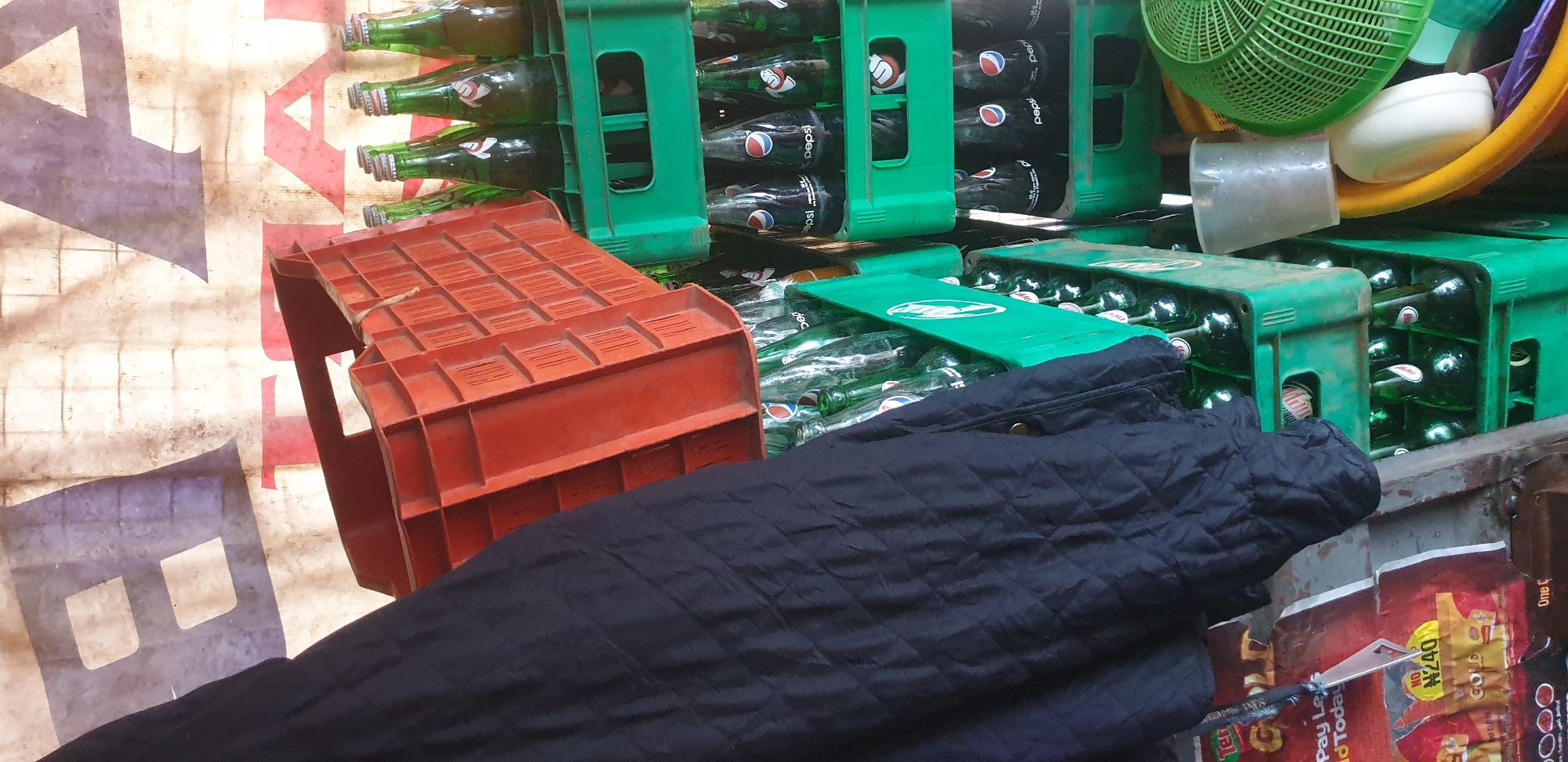 A close-up, vertically oriented photograph captures stacks of green plastic beverage crates filled with glass bottles, predominantly Pepsi, in what appears to be an outdoor market stall. An orange plastic crate is partially visible in the upper left, tilted and resting on top of the green crates. To the left of the crates, a dark blue quilted jacket is draped over an unseen surface, obscuring a portion of the background. The background is a beige tarp or canvas with large, faded blue and red lettering, suggestive of signage.
Several layers of green crates are stacked, each holding multiple rows of green glass bottles. The bottles are of varying fill levels, with some appearing full and others partially empty. The Pepsi logo is clearly visible on many of the bottles. Some bottles have silver caps, while others have darker colored caps.
Below the crates, to the left, is a portion of a promotional poster or flyer. It features yellow and red colors and text including "GOLD", "Pay Less", and "N240".
To the right of the stacked crates, a clear plastic cup filled with ice or small pebbles is positioned next to a large, bright green plastic basket, possibly for carrying produce. A yellow basin or bowl is also visible in the lower right corner, containing a white, oval object that resembles soap or a bar of detergent.
The lighting suggests an outdoor setting, likely during the day, with some shadows indicating the direction of the light source. The overall impression is one of a bustling market environment, possibly a point of sale for beverages and other goods. The scene depicts an inventory of drinks ready for sale.
