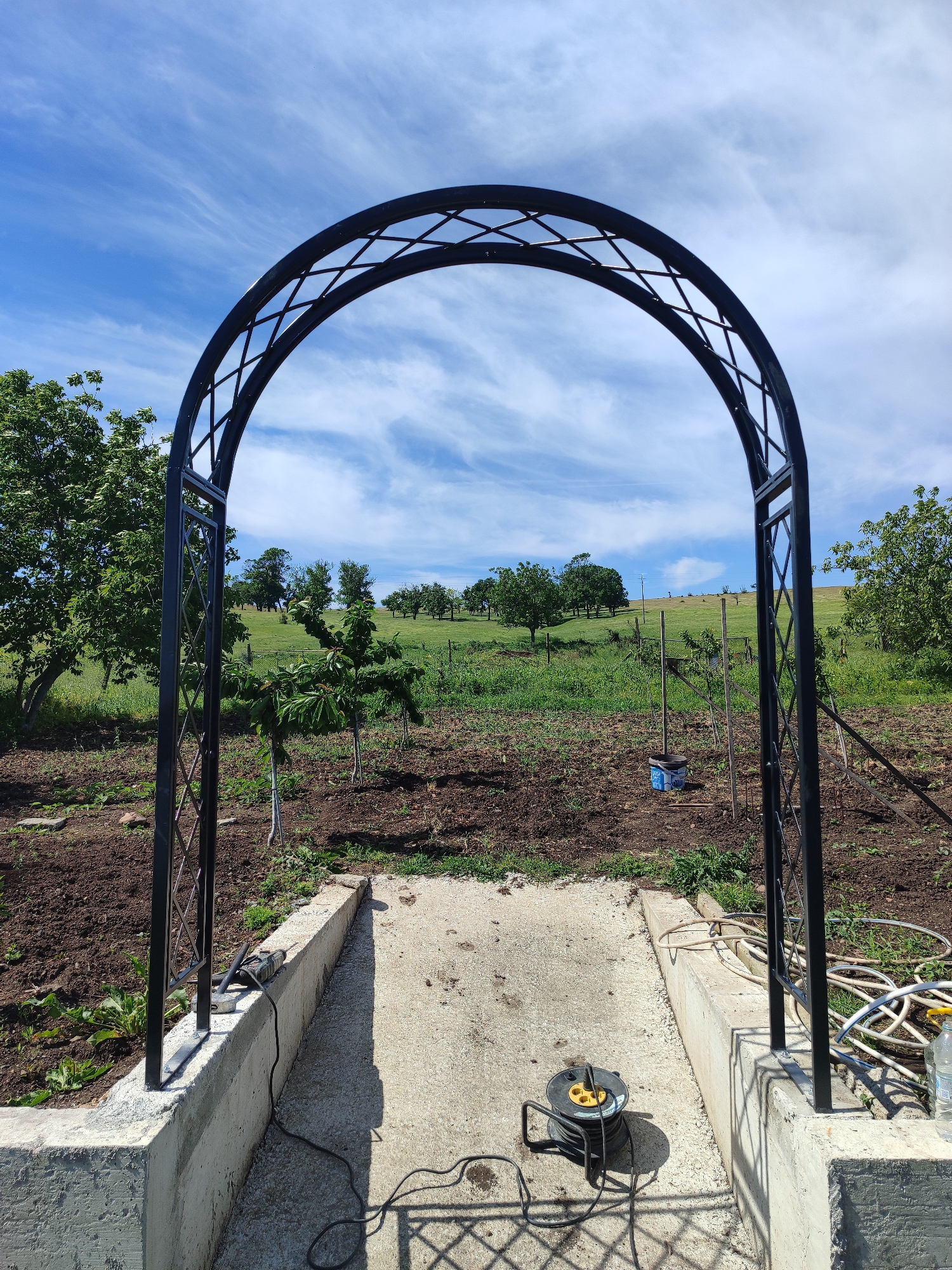The image shows a black metal garden archway erected on a concrete platform in Bata, Bulgaria. The archway frames a view of a cultivated garden with young trees and a grassy hillside in the background under a bright blue sky with scattered clouds. An electrical cord reel sits on the concrete platform in the foreground, with tools, a water bottle, and garden hoses nearby, suggesting ongoing construction or landscaping work.