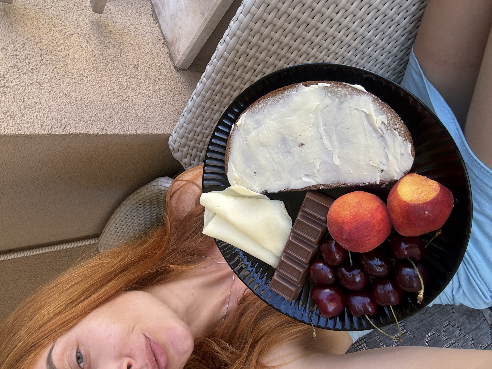 In Budva, Montenegro, a woman with striking red hair is seated outdoors, holding a black plate laden with a breakfast spread. The plate contains a slice of dark bread generously buttered, a bar of milk chocolate, several ripe cherries, two peaches, and slices of cheese. The woman is wearing a light blue top. The background reveals a textured, light brown wall and a portion of a woven outdoor chair, suggesting a relaxed, alfresco setting, possibly a balcony or patio. The overall impression is one of a pleasant morning meal enjoyed in an outdoor environment.