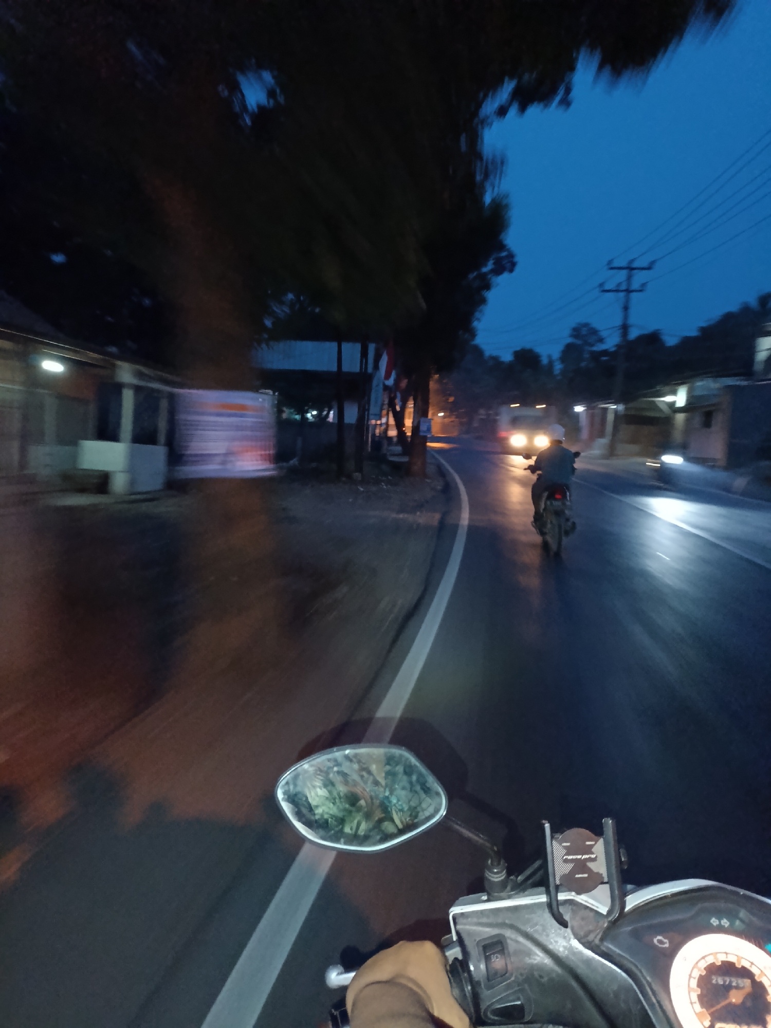 A first-person perspective, low-light image captures a motorcycle's dashboard and a rear-view mirror while the vehicle is in motion on a wet asphalt road. The rider's left hand, wearing a light-colored sleeve, is visible near the handlebars. Ahead, another motorcyclist wearing a helmet and dark jacket is riding away from the camera, illuminated by the headlights of an approaching vehicle. The scene appears to be a street in Jonggol, Indonesia, at dusk or night, with sparse streetlights and building facades visible on the sides of the road. Trees and utility poles line the path. The road surface is reflective, indicating recent rain or dampness. A phone mount is attached to the motorcycle's dashboard. The dashboard display shows a digital odometer reading of "26726."
