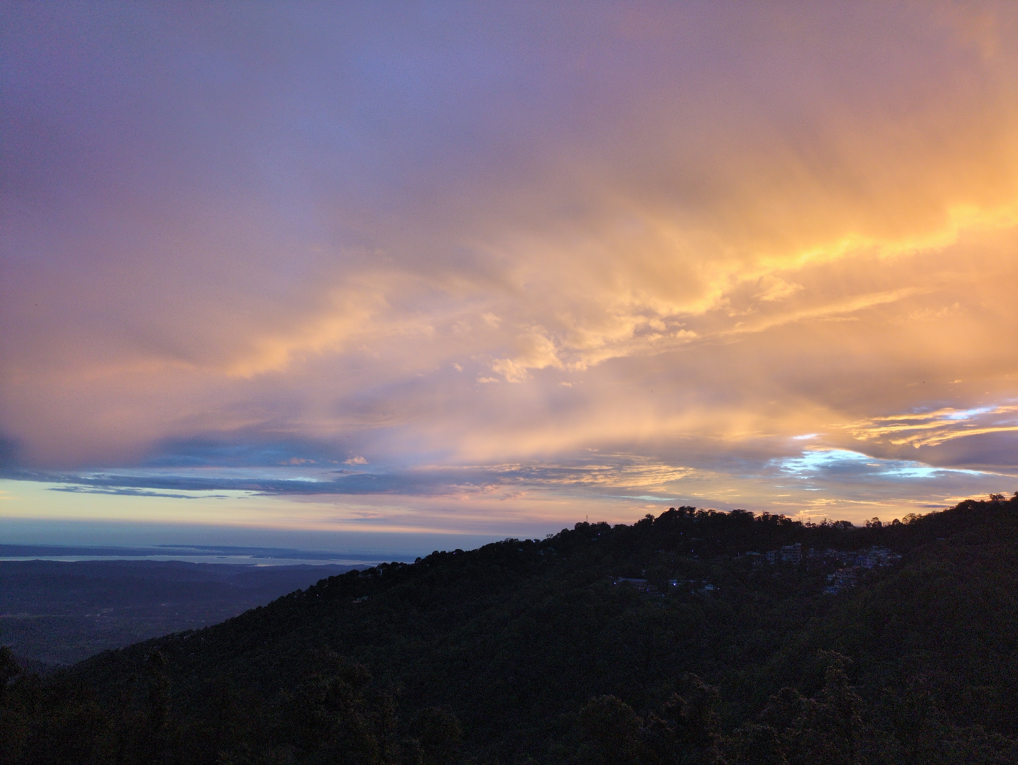 The image captures a twilight scene over a mountainous landscape, likely in or near Dharamkot, India, given the context. The sky is filled with dramatic clouds painted in hues of purple, pink, and vibrant orange from the setting sun. The upper portion of the sky is a soft lavender, transitioning to more intense colors as it meets the clouds. A bright band of golden light illuminates the edges of some of the clouds, suggesting the sun is just below the horizon. Below the sky, a dark, densely forested hillside dominates the foreground and midground. The silhouette of the trees creates a rich, textured mass that slopes downward from right to left. Scattered amongst the trees on the hillside, particularly along the ridge, are small clusters of buildings, indicated by faint lights, suggesting a small settlement or village. In the far distance, a body of water, possibly a lake or bay, is visible, bordered by a hazy, lighter horizon. The overall atmosphere is serene and expansive, with the natural beauty of the sky and landscape taking center stage. There are no visible people or discernible actions taking place.