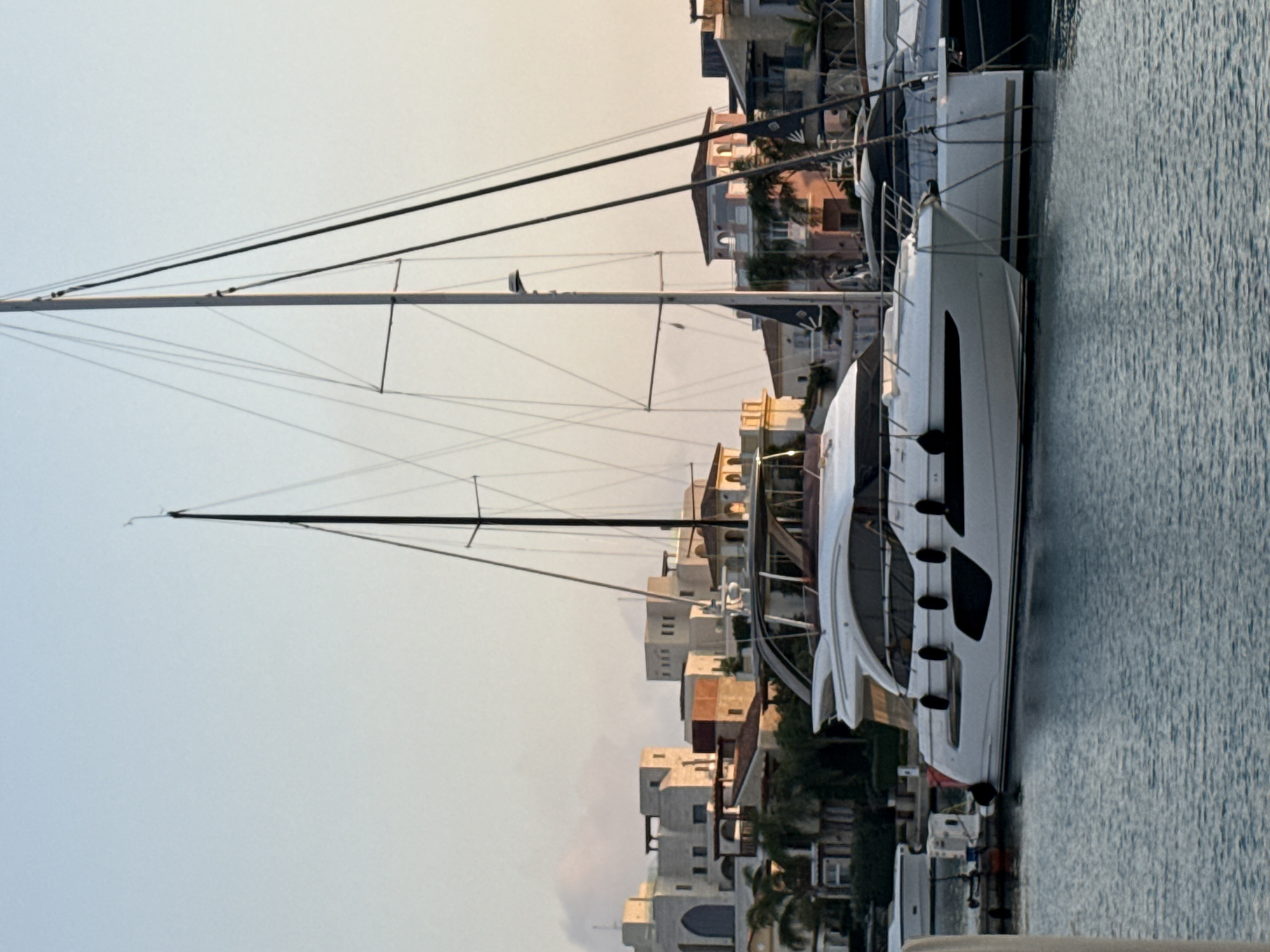 In Limassol, Cyprus, a luxury white yacht is moored in a marina. The yacht features sleek lines and dark accents, with several black fenders attached to its port side. Behind the yacht, a cluster of buildings with Mediterranean-style architecture, characterized by light-colored facades and flat roofs, are visible. These buildings are interspersed with palm trees, suggesting a warm climate. Two sailboats with tall masts stand in the background, their rigging creating a complex network of lines against the clear, pale blue sky. The water in the marina is calm and reflects the sky and boats. The scene appears to be captured during a sunset or sunrise, indicated by the soft, warm light illuminating the buildings and the sky.