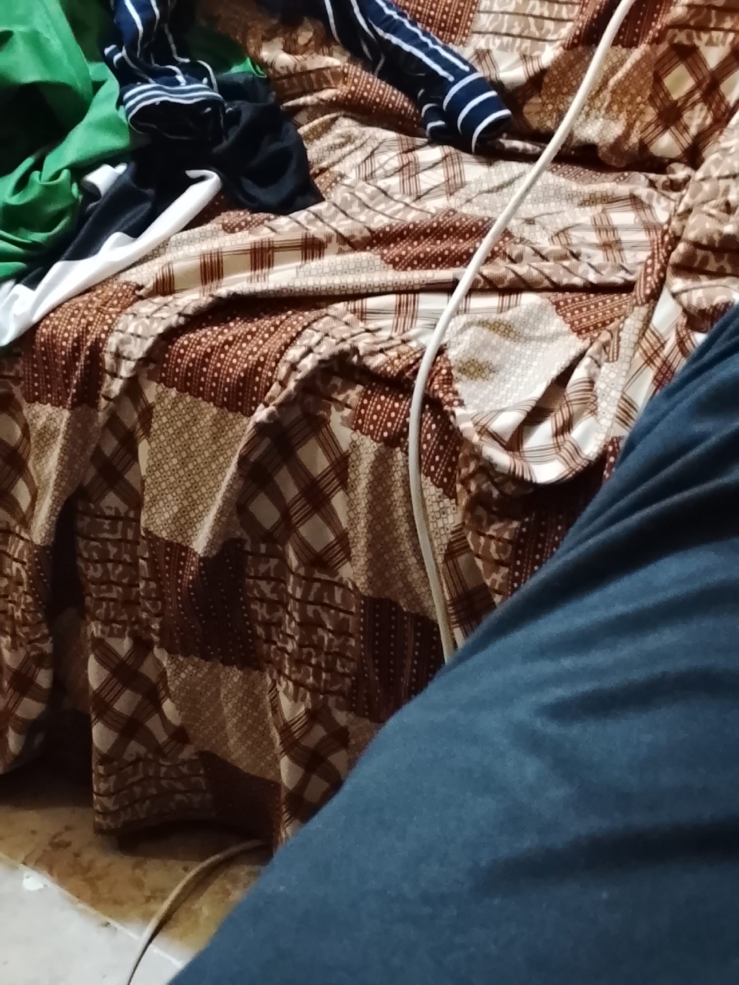 A medium shot shows a portion of a patterned bedcover with various clothing items atop it. Green, white, and blue striped garments are among the items. A white electrical cord is visible on the bedcover. A large expanse of dark blue fabric occupies the lower right foreground, possibly clothing. The image was taken inside a room. The location is Multan, Pakistan.
