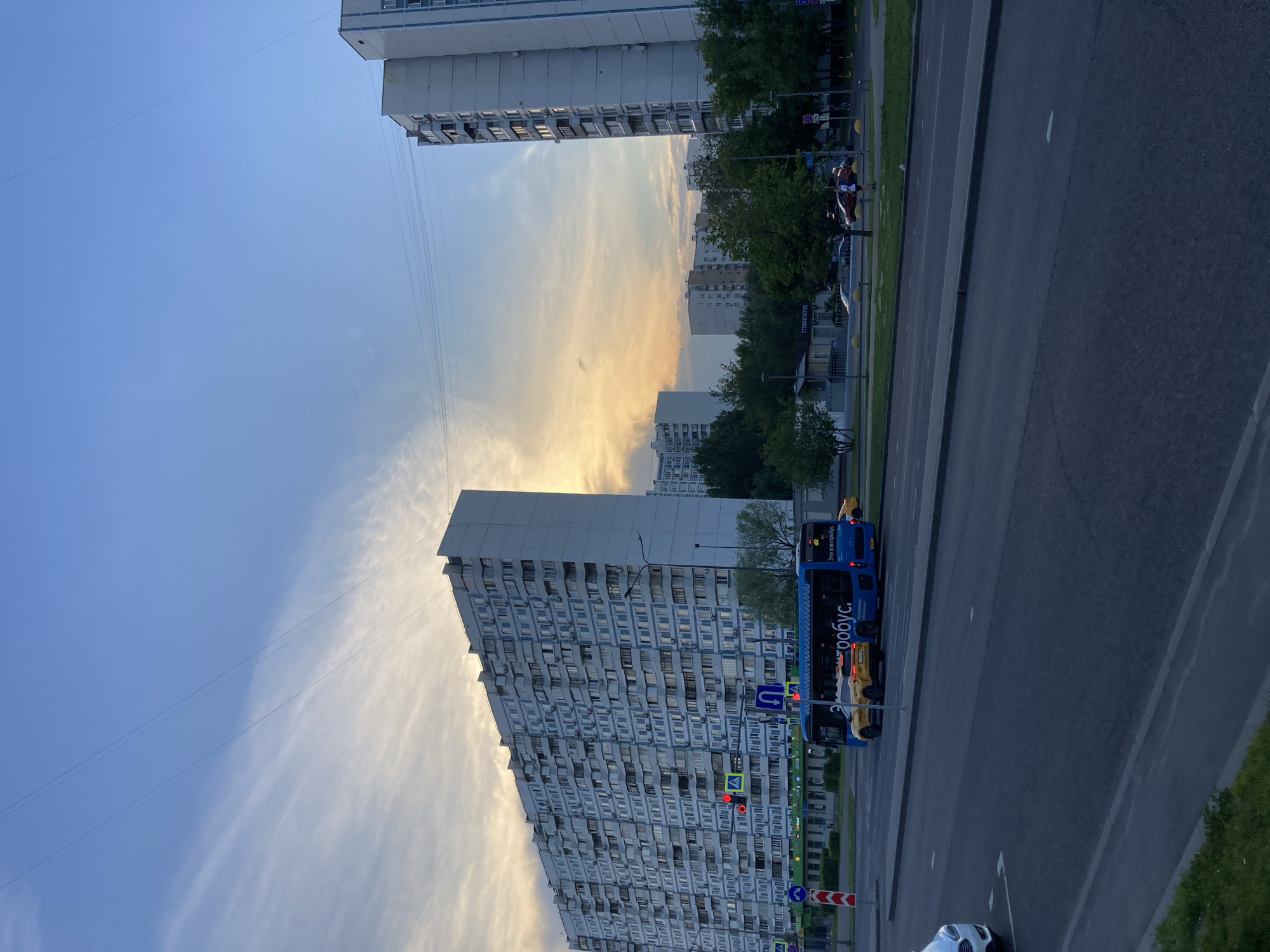 This is a photograph taken at dusk, capturing a wide view of an urban street scene with high-rise apartment buildings and a roadway in Moscow, Russia. The sky is a soft blue, transitioning to a warm yellow and orange hue near the horizon, indicative of sunset, with wispy clouds present. In the foreground, a paved road with lane markings is visible, with a white car on the left. A blue electric bus, labeled with Russian text, is driving on the road, and next to it is a yellow taxi. Behind the road, a row of trees lines the sidewalk, with a pedestrian walkway. Several buildings are visible, including a very long, multi-story apartment building on the left with many windows and balconies, and a taller apartment building on the right. In the background, other apartment buildings and trees are scattered across the landscape. Traffic lights and street signs are also present.