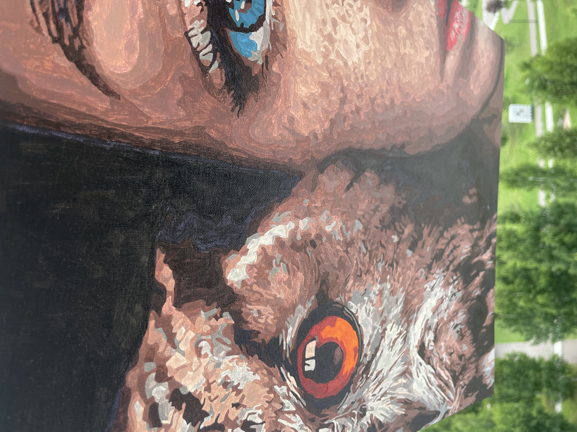 A close-up, detailed oil painting is displayed, featuring a split image of a human face and an owl's face. The human eye is rendered in vibrant blue, with dark eyelashes and a fair complexion. To the left, the owl's eye is a striking orange, with visible pupil and surrounding feathers depicted in shades of brown and white. The background of the painting is a dark, textured surface. The painting is oriented horizontally and is propped up against a railing, with a blurred background of green trees and a pathway visible below. The setting appears to be outdoors, possibly a balcony or elevated vantage point in a park or residential area.