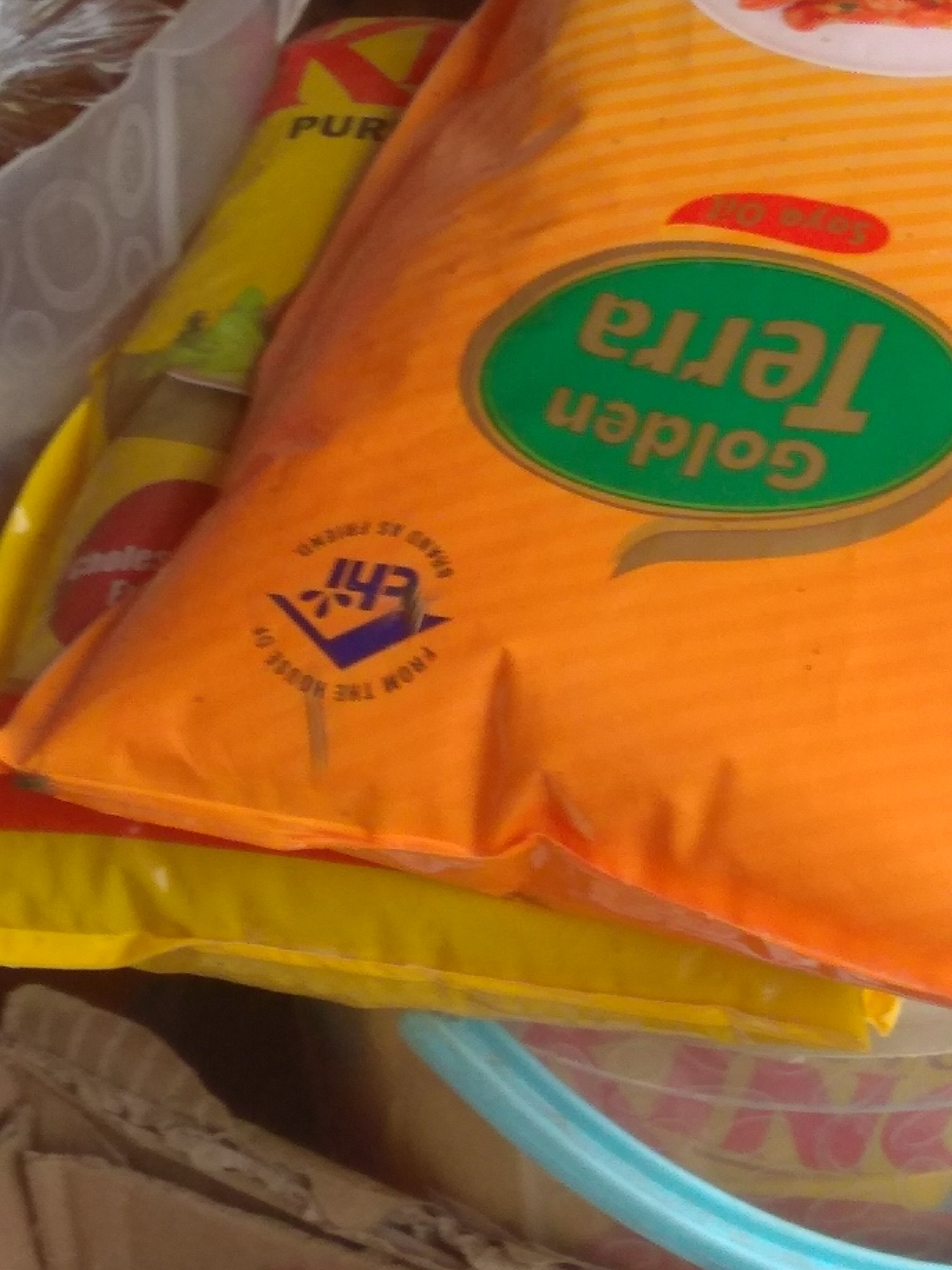 The image shows a close-up, slightly blurry view of stacked packaging. The most prominent item is a large, bright orange bag with a green oval logo containing gold lettering that reads "Golden Terra" in a curved, downward orientation. Above this logo, a red oval shape with white text is partially visible, appearing to say "Soro Oil" or similar. On the lower part of the orange bag, there is a circular emblem with blue and white text. The text on this emblem is difficult to read clearly, but appears to include "FROM THE HOUSE OF" and possibly an abbreviation like "EFI" or "EHI" in the center.
Behind the orange bag, to the left, is a yellow package with red lettering that partially spells out "PUR". Further behind this, a white translucent bag or material is visible. Below the orange and yellow packaging, another layer of yellow packaging is seen, and beneath that, the rim of a blue plastic bucket is visible, with some red and pink lettering on the bucket's contents.
The image is taken indoors, with artificial lighting. There are no people or discernible outdoor elements visible. The location in Jalingo, Nigeria, is not evident from the visual information within the image itself.