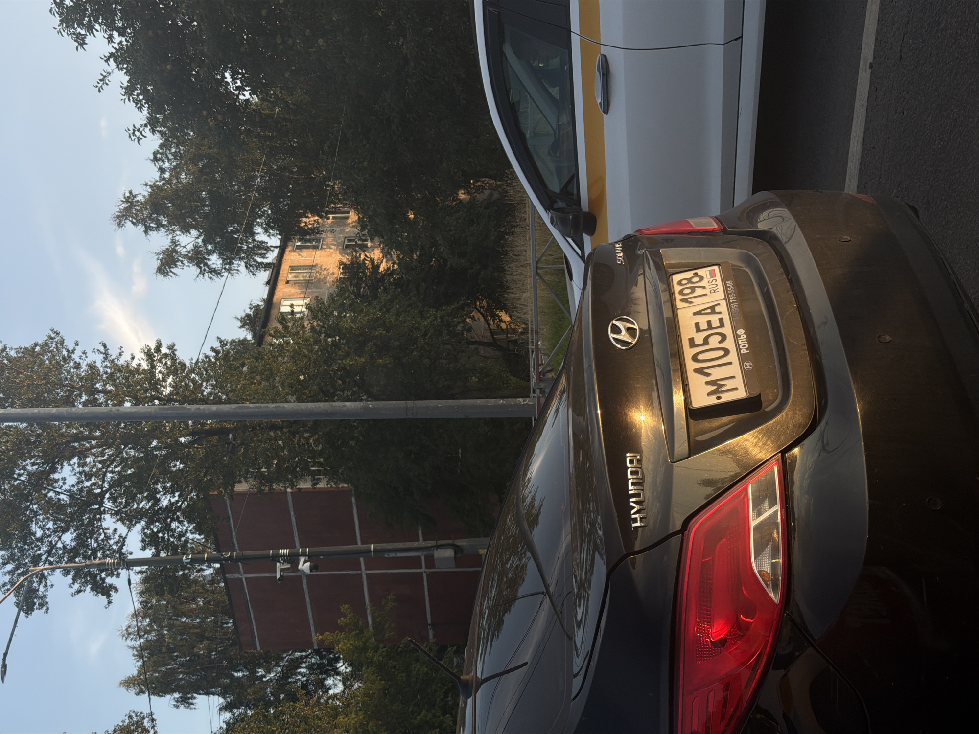 The image captures a street scene in Saint Petersburg, Russia, likely during late afternoon or early evening given the warm, golden light. In the foreground, the rear of a dark Hyundai sedan is prominently featured, its taillight and "HYUNDAI" badging illuminated by the setting sun. The license plate is visible, displaying the characters "M105EA 198" and "RUS", indicating it is a Russian vehicle. To the right, a white car, possibly a taxi due to the prominent yellow stripe along its side, is parked next to the Hyundai. In the background, a multi-story residential building with visible windows stands amongst lush green trees. A lamppost with what appears to be security cameras attached is also visible, along with other power lines and foliage. The overall impression is a typical urban environment in Russia, with cars parked on a street bordered by buildings and trees.