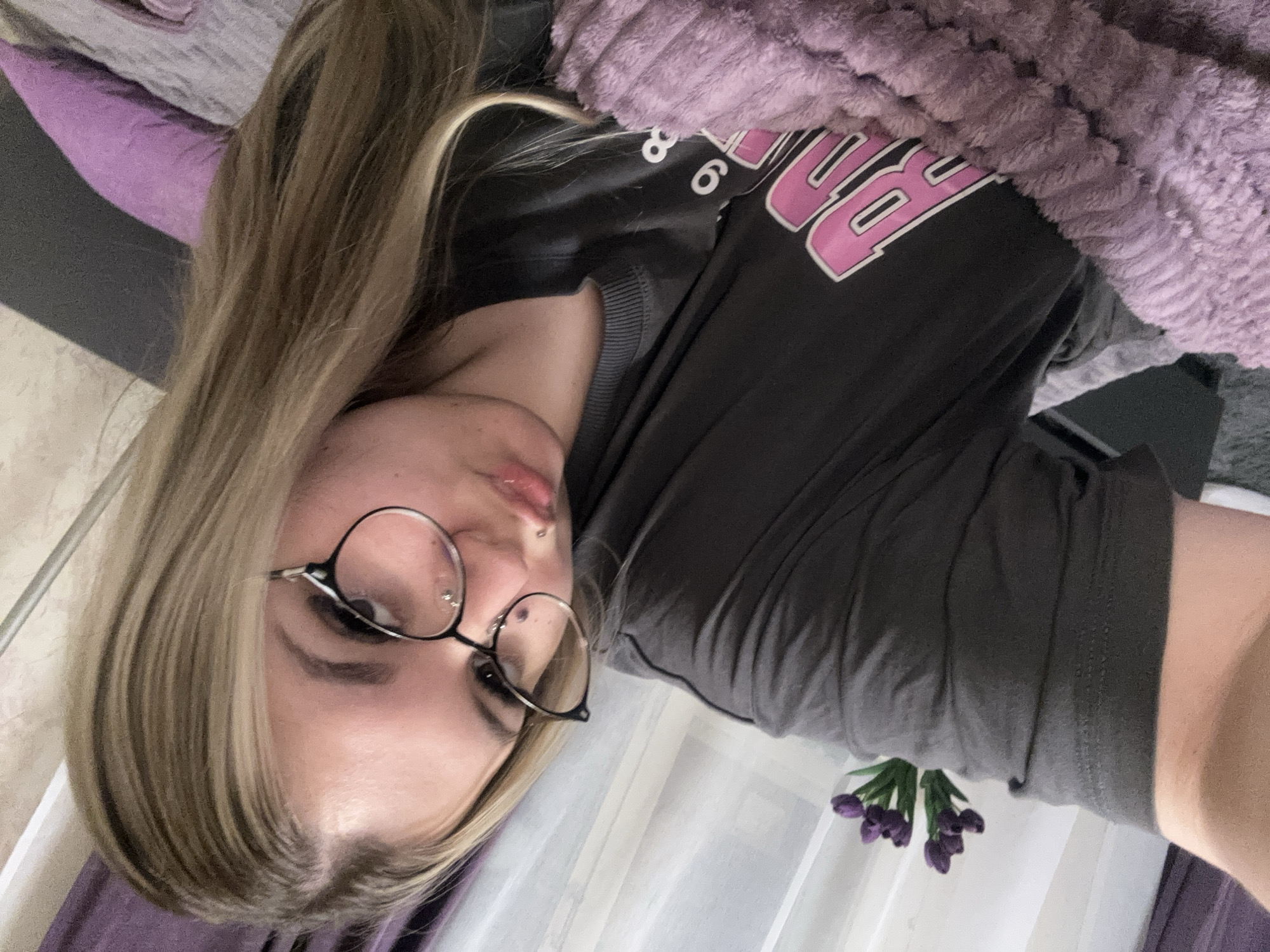 A young woman with long, straight blonde hair and darker roots or highlights is depicted in a close-up, appearing to be indoors and possibly reclining. She wears round, black-framed glasses and has a philtrum lip piercing. Her expression is neutral. She is dressed in a dark grey t-shirt featuring a prominent pink graphic design, over which a textured, fluffy purple blanket is draped across her shoulder and upper body. The t-shirt's graphic includes partially visible stylized pink capital letters with white outlines, notably the numbers "8" and "6". In the foreground, to the right, a cluster of small purple flowers, possibly artificial tulips, rests on a white surface with a sheer white curtain behind them. A purple pillow or fabric is visible on the left side of the frame. The scene is well-lit, suggesting daytime indoors. No specific outdoor weather or geographical cues for Большой Букор, Russia, are discernible from the image.
