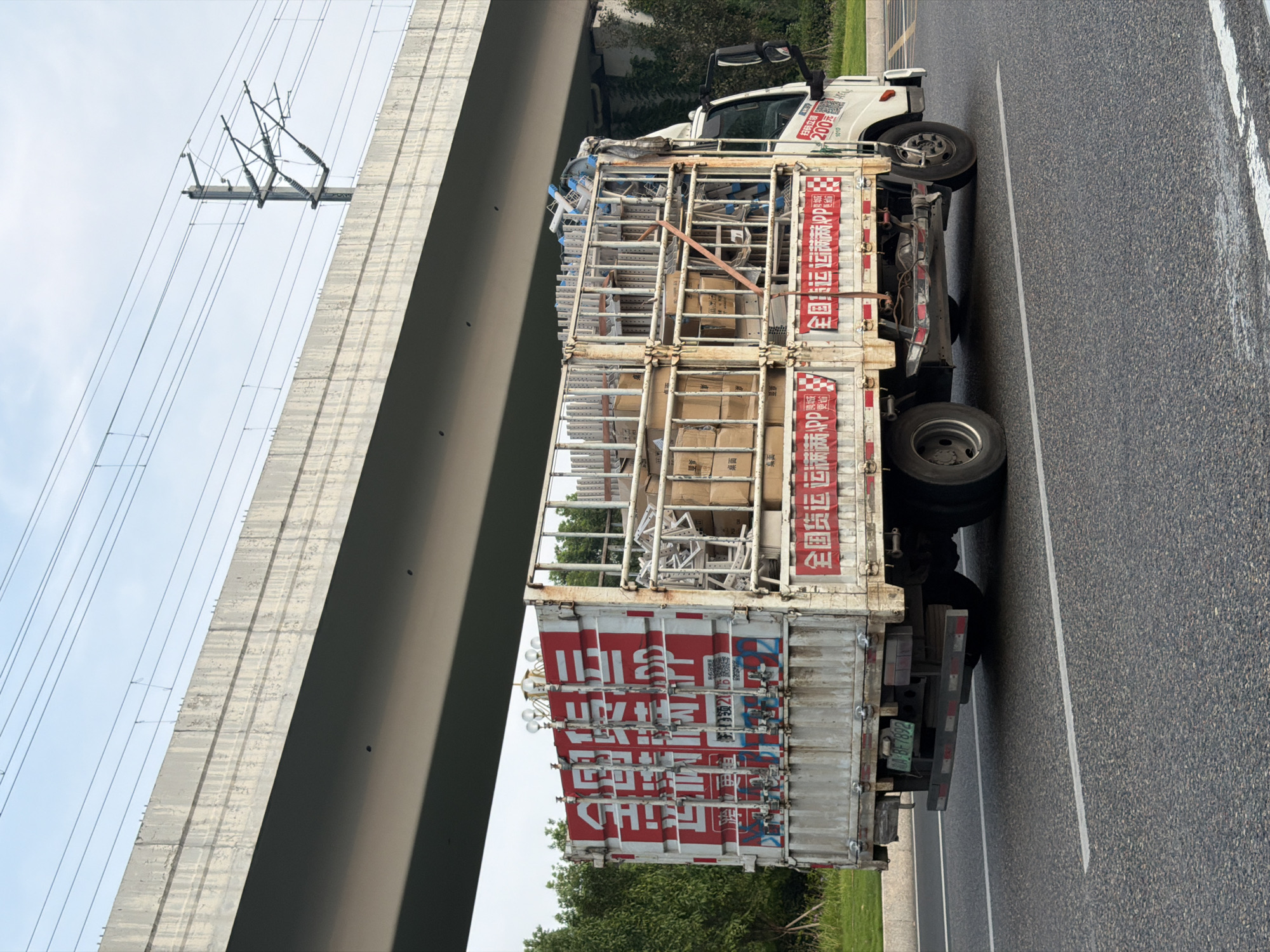 In the city of Jiangjin, China, a white flatbed truck with a metal cargo cage is driving on an asphalt road. The truck is loaded with boxes and metal shelving units. The side of the cargo cage is decorated with large red and white Chinese characters that read "全国货运APP" (National Freight APP). The truck has a license plate that reads "渝BF7892". In the background, there is a large concrete overpass and overhead power lines. The sky is cloudy.