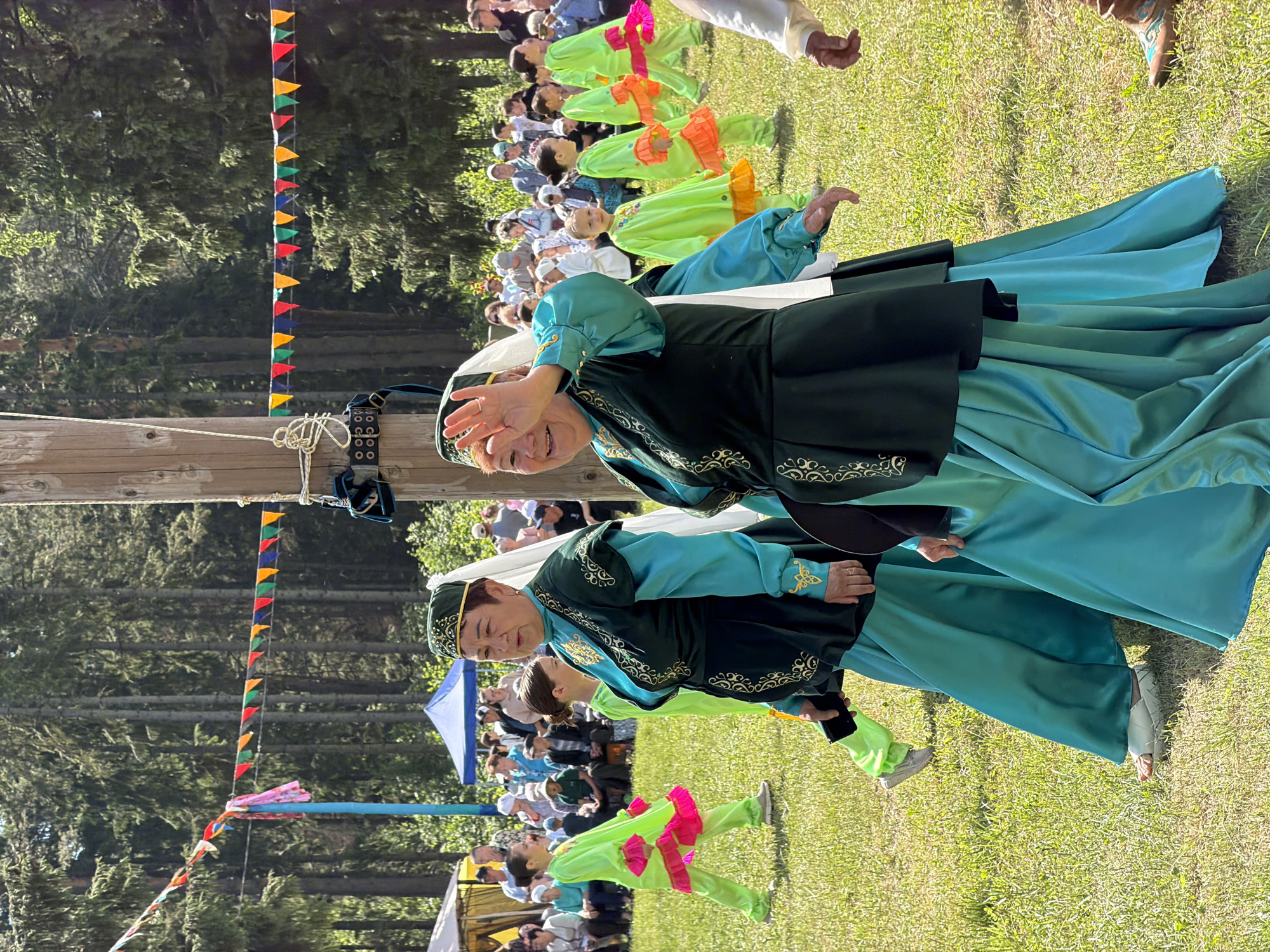 An outdoor scene depicts a cultural event on a bright, sunny day within a mown grassy field. A dense forest of tall evergreen trees forms the background.
In the foreground, two middle-aged to older women, dressed in matching traditional-style attire, walk from left to right. Their outfits consist of a dark green over-tunic with intricate gold embroidery, worn over a long-sleeved, flowing teal dress. Each woman wears a dark green cap with gold trim and a white veil or scarf draped from the back of the headpiece. The woman on the left holds a black smartphone in her left hand. The woman on the right is smiling and waves her right hand forward; a ring is visible on her left ring finger. She wears white platform sandals.
Behind the two women, several children, primarily girls, walk in a line, also moving from left to right. They are dressed in bright lime-green outfits with contrasting ruffled trim; some have hot pink trim, others have bright orange trim on their sleeves and pant cuffs or skirt hems.
In the far background, a large crowd of spectators, some seated under canopies or tents (one beige, one blue), observe the event.
A tall, unpainted wooden pole stands vertically on the right side of the frame, adorned with ropes and a black harness-like object with silver buckles. A string of colorful triangular bunting, featuring red, yellow, green, and blue flags, stretches across the upper mid-ground, attached to the wooden pole and another light blue pole on the left side of the frame. A partial view of a person's leg wearing a brown boot with light blue embroidery is visible at the far bottom right.
