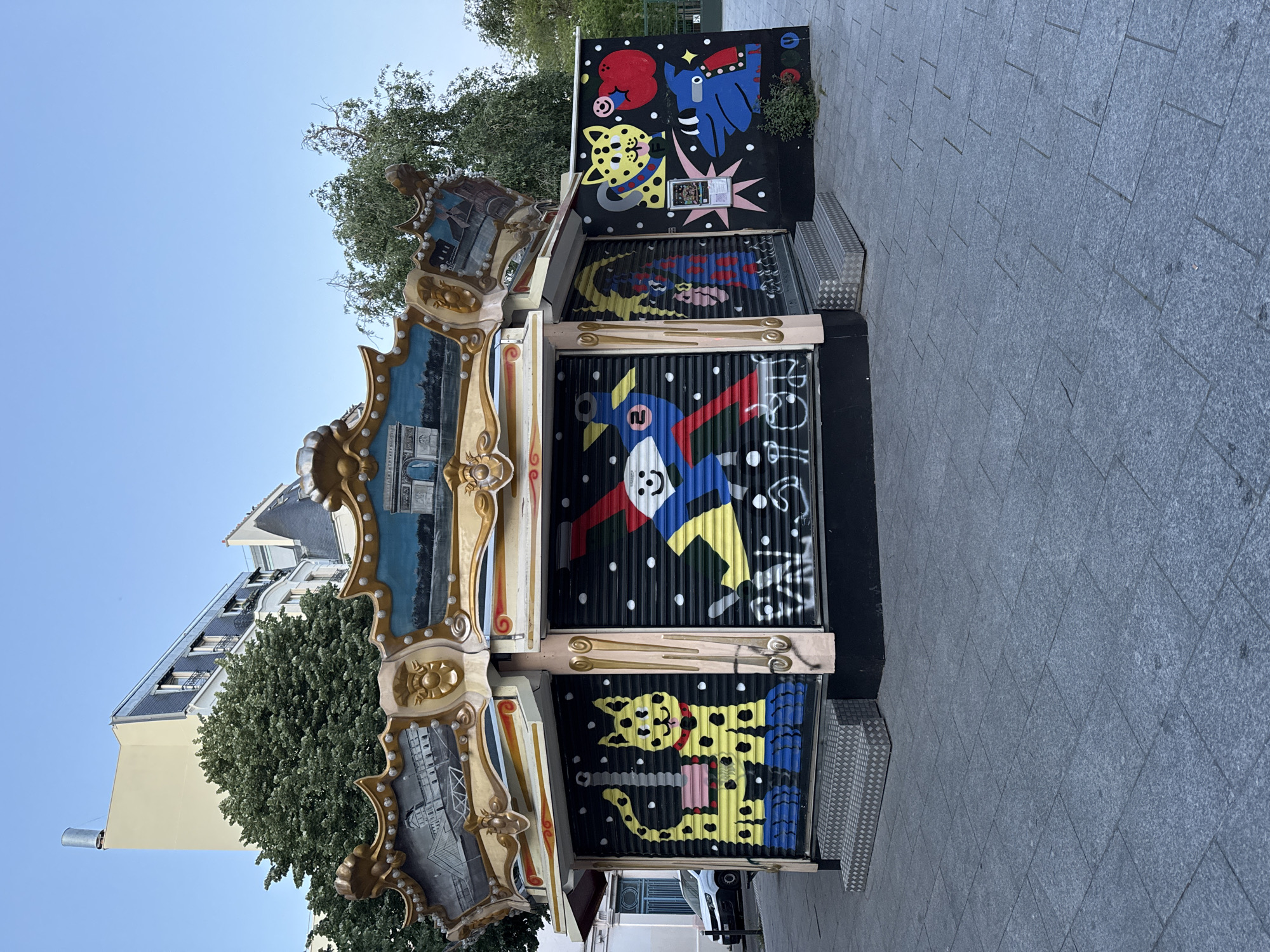 An upright outdoor scene features a vibrant, closed carousel or kiosk adorned with both traditional and modern artistic elements, set against a backdrop of urban architecture and a clear blue sky.
The central subject is a brightly painted structure. The upper part displays classic carousel aesthetics with gilded, ornate frames surrounding painted panels, depicting what appears to be Parisian landmarks like the Arc de Triomphe and a river scene. Below these, the kiosk is secured by corrugated metal roller shutters, which are covered in striking, cartoon-like graffiti. The dominant color scheme for the graffiti is black background with white dots, featuring yellow spotted cat-like creatures, a blue dog-like figure with a red heart, and a blue and yellow bird-like character, all with bold outlines and contrasting colors like red and green. Some white graffiti text is visible on one of the shutters, but it is largely illegible. On the top right panel of the kiosk, a small framed sign is visible, but its text is unreadable. To the right of the kiosk, on a black wall, a red circular logo with a blue bar, resembling the London Underground symbol, and a stylized "M" (likely for Métro) are present.
To the right of the kiosk, a clean, grey paved sidewalk extends into the distance, composed of rectangular stone tiles. A textured metal utility box or bench sits on the sidewalk near the kiosk. In the background, lush green trees partially obscure multi-story buildings, characteristic of Parisian architecture, featuring light-colored facades and dark roofs. The sky is clear and light blue, suggesting good weather during the day, possibly morning or afternoon, given the soft lighting and lack of harsh shadows. The kiosk appears to be closed, indicating no immediate activity or events are taking place, and no people are visible in the scene.