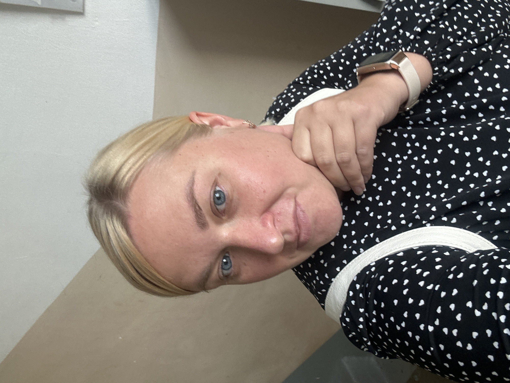 The image shows a selfie of a blonde woman with blue eyes, wearing a black dress with a white heart pattern and a light-colored strap across her shoulder. She is resting her chin on her hand and looking at the camera. A smartwatch is visible on her wrist. The background appears to be an indoor setting with a wall that transitions from beige to white, suggesting a neutral and possibly unfinished or minimalist interior design. The lighting is natural and bright, illuminating the woman's face and features. There are no specific identifiable landmarks or elements that would indicate a location in the city of Chaikovsky, Russia.