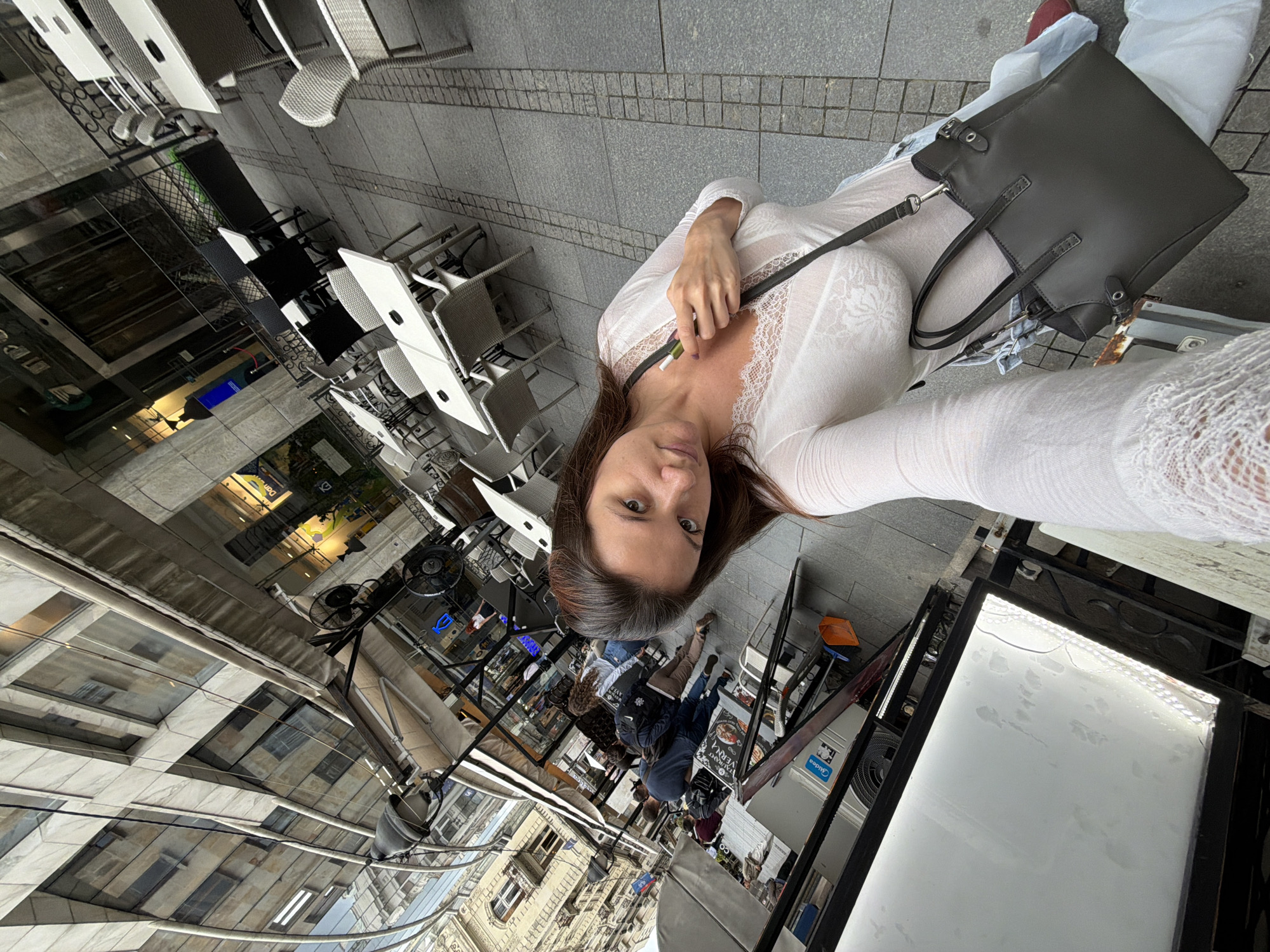 The image depicts a woman taking a selfie while standing on a street in Belgrade, Serbia. She is wearing a white, long-sleeved, lace-trimmed top, jeans, and a gray tote bag. She holds a vaping pen. The background shows outdoor seating areas of cafes and restaurants with tables and chairs along a pedestrian street. Pedestrians can be seen walking in the distance. The architecture suggests a historic urban area. The sky is overcast, casting a soft light over the scene.