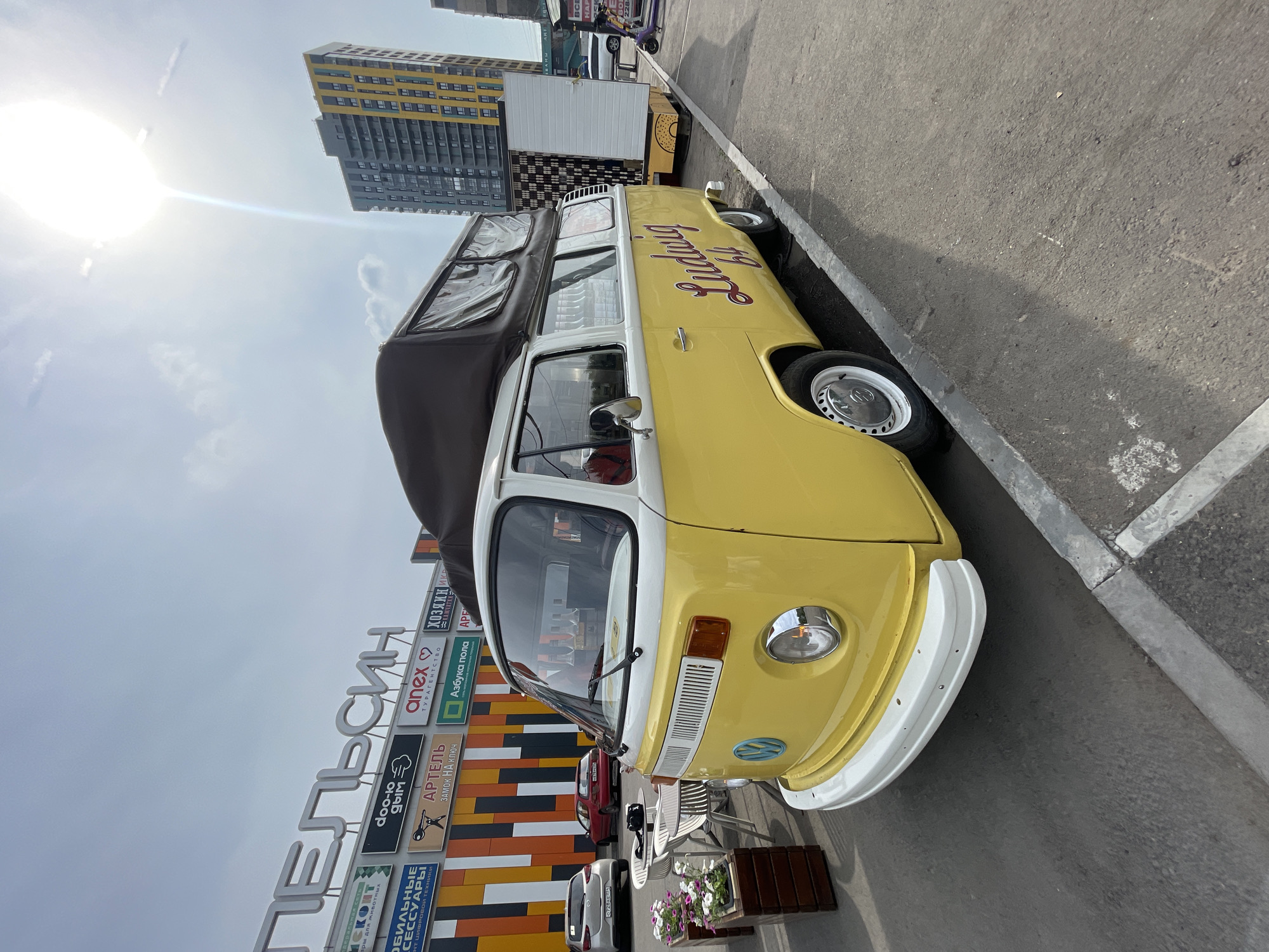 This is a wide-angle outdoor photograph taken on a sunny day in the city of Krasnoyarsk, Russia. The primary subject is a yellow and white Volkswagen Type 2 camper van parked on an asphalt surface. The van has a pop-up tent on its roof and the word "Ludwig 64" is written in red script on its side. To the left of the van, there are several white tables and chairs set up, along with planters containing pink and white flowers. Behind the van and tables is a building with a large sign that reads "ПЕЛЬСИН" (PELSIN) in large white letters. Beneath this main sign are several smaller signs for various businesses, including "doo-lo grim", "anex", "АРТЕЛЬ ЗАМОК НА КЛЮЧ", and "АЗБУКА ПОЛА". To the right of the van, there is a white food truck or kiosk with a yellow donut design on its side. In the background, a modern, multi-story building with yellow and grey facades is visible, along with other less distinct buildings and some electrical wires. The sky is mostly clear with a few scattered clouds, and the sun is bright, creating a lens flare effect in the upper right quadrant of the image. There are no people clearly visible in the photograph. The scene appears to be an outdoor seating area or market setup next to a commercial building.