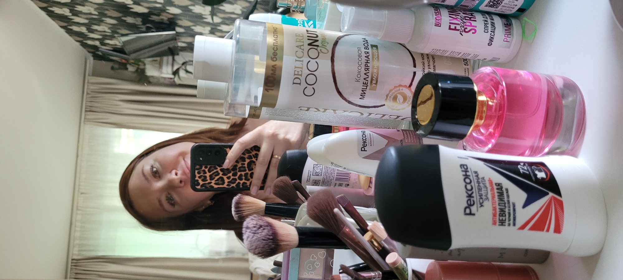 The image is a selfie taken in front of a mirror. The reflection shows a woman holding a smartphone with a leopard-print case. A variety of cosmetic products are arranged in front of her on a surface, including bottles of "Delicare Coconut" micellar water, "Rexona" deodorant, a pink perfume bottle, and "Luxvisage Fixing make-up Spray". Also present are makeup brushes. The background visible in the mirror shows part of a room with a patterned curtain and a wall. The location of the selfie is Unknown, Unknown.