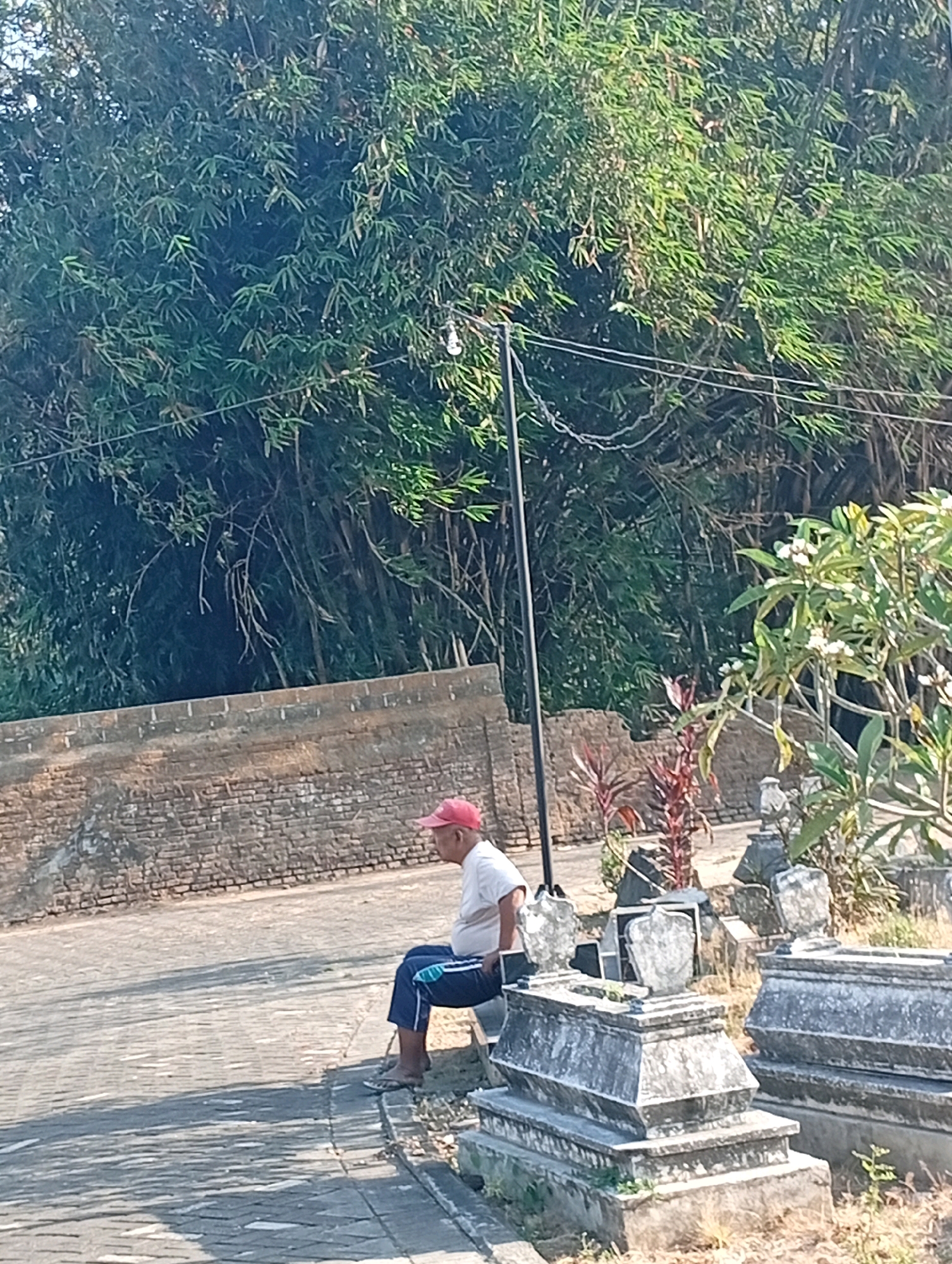 A man wearing a red cap, a light-colored short-sleeved shirt, and dark blue pants with a light blue stripe sits on a bench facing right in what appears to be a graveyard. The man is positioned in the lower right quadrant of the image, near a row of stone tombs. Behind him, a brick wall runs horizontally across the middle ground, topped with lush green bamboo trees that dominate the upper portion of the frame. The ground in front of the man and tombs is paved with interlocking stones, with a shadow cast across it. The scene is lit by bright sunlight, suggesting it is daytime. The image is taken from a medium distance, capturing the man, the tombs, the wall, and the bamboo foliage. The overall setting suggests a peaceful, possibly contemplative atmosphere in a historical or cultural site.