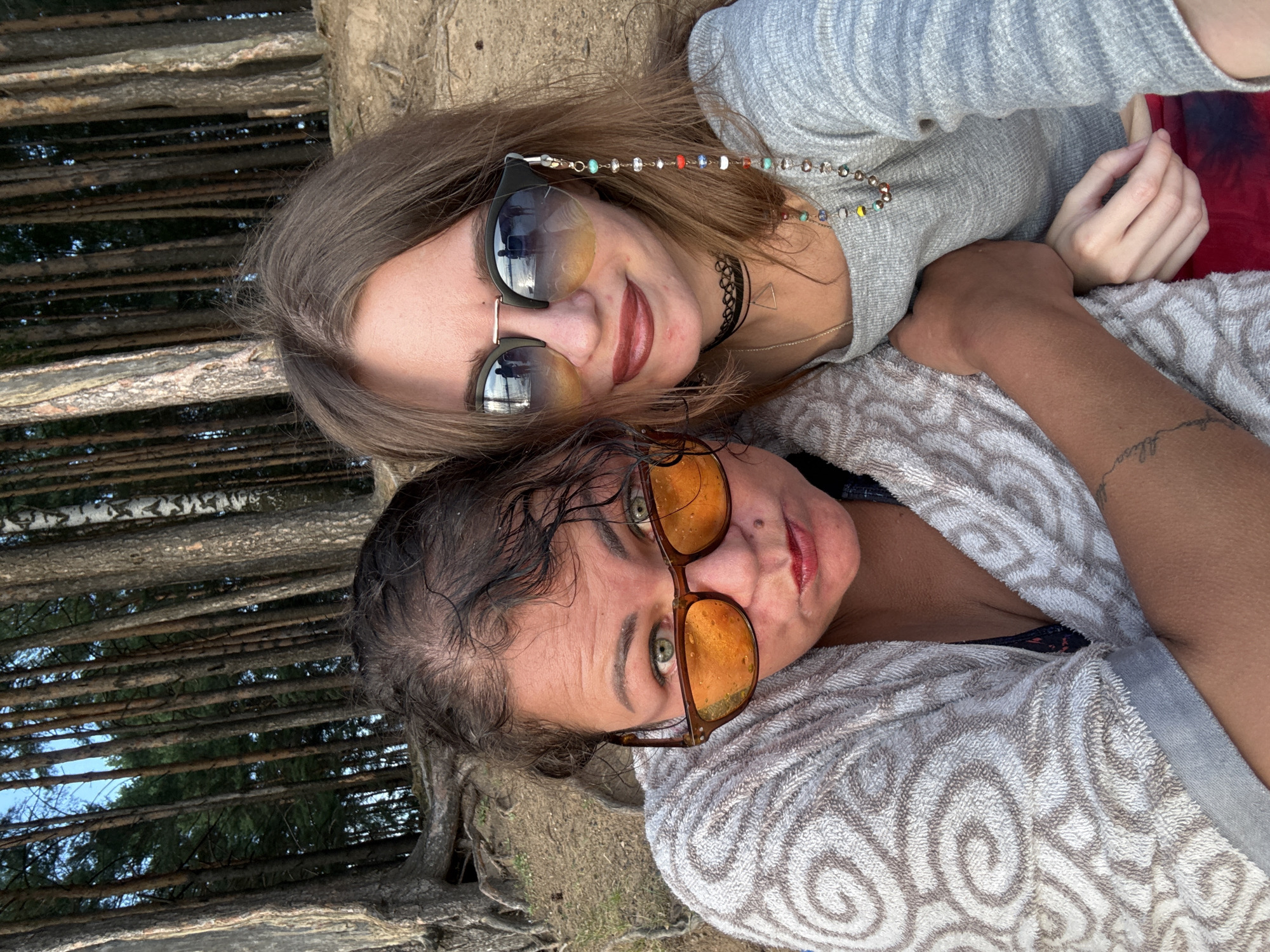 Two women are taking a selfie in a forest with tall trees and a sandy ground. The woman on the left has dark curly hair and is wearing a grey patterned robe over a swimsuit. She has on large, amber-tinted sunglasses and red lipstick. The woman on the right has long, light brown hair and is wearing a grey long-sleeved shirt, a black choker necklace, and sunglasses with a beaded strap. They are positioned close together, with the woman on the left's arm around the woman on the right. The background consists of a dense forest with thin, tall trees and some exposed roots. The ground appears to be sand and dirt.