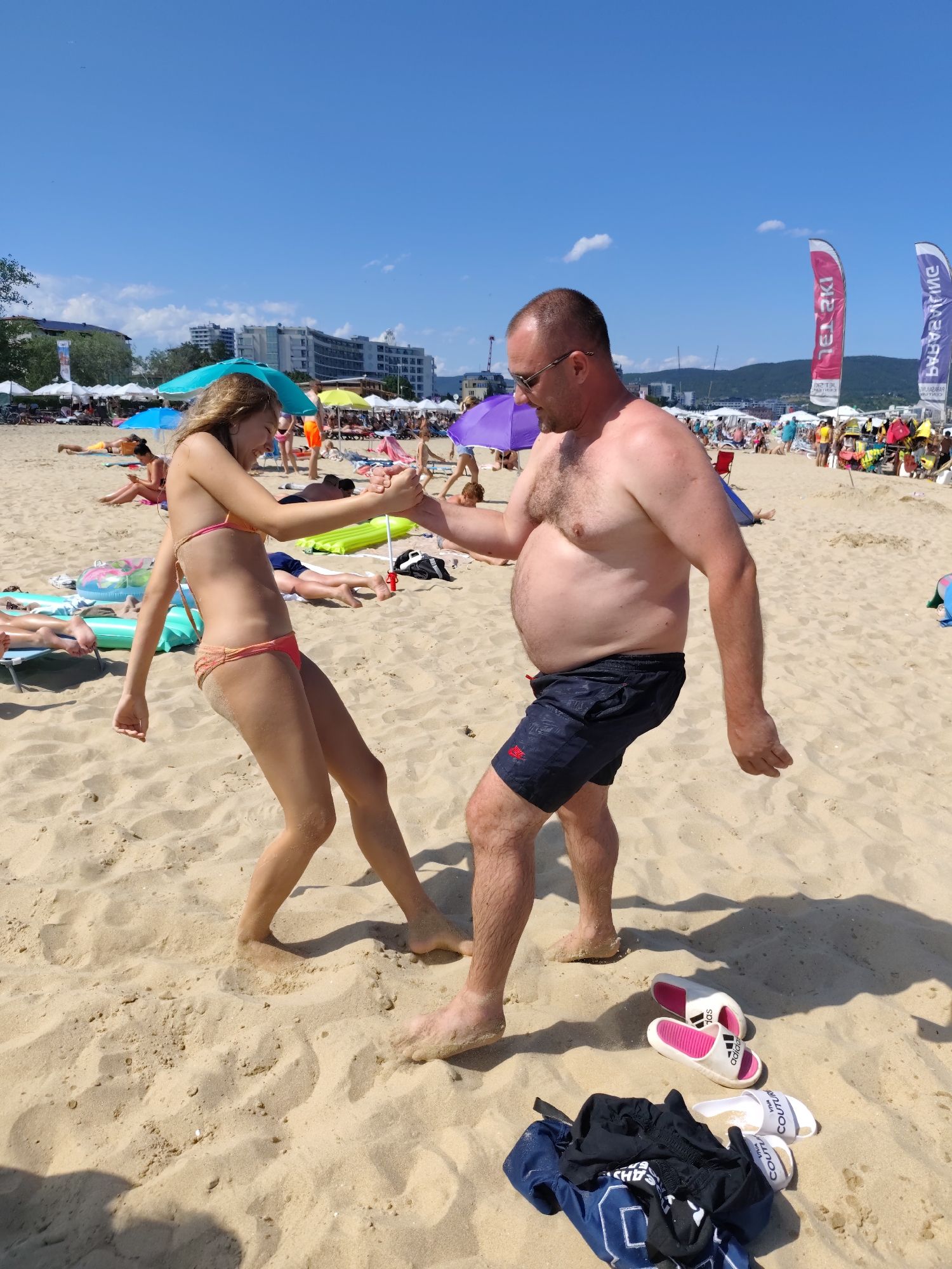A man and a young woman are dancing on a sandy beach. The man, wearing sunglasses and black swim shorts, has his hand clasped with the woman's. She is wearing a coral bikini. Both appear to be enjoying themselves, with the woman smiling.
The scene is a crowded beach with many other people sunbathing and relaxing on lounge chairs under umbrellas. The background shows buildings and hills, suggesting an urban coastline. The sky is bright blue with scattered clouds, indicating a sunny day, likely midday given the strong shadows. The presence of beach umbrellas, lounge chairs, and swimwear indicates a leisure activity.
In the foreground, to the right of the man, a pair of white Adidas slides with pink accents and another pair of white slides with "COUTUR" visible are placed on the sand next to a pile of dark clothing. In the background, two tall, pink and white flags with text are planted in the sand. One flag has Cyrillic text that reads "НЕСЕБЪР" (Nesebar). This, along with the visual cues of the buildings and landscape, confirms the location as Nesebar, Bulgaria. The overall atmosphere is one of a vibrant, summer beach day.