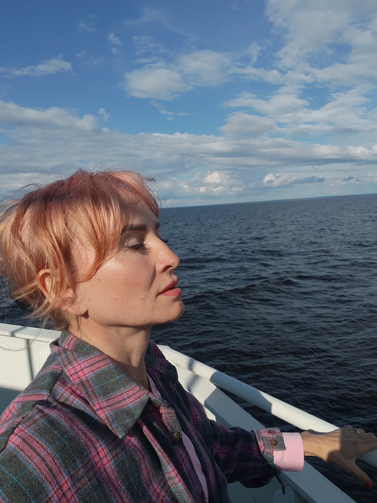 A medium shot captures a woman in profile, standing on what appears to be a boat deck. Her head is tilted upwards and her eyes are closed, as if enjoying a moment of reflection or basking in the sun. Her short, peachy-pink hair is slightly windswept. She wears a pink and gray plaid shirt over a light pink garment. Her left hand rests on a white railing that runs along the edge of the deck. The deep blue water of a large body of water stretches out behind her, meeting a horizon line under a bright blue sky dotted with white, fluffy clouds. The lighting suggests it is daytime, likely late afternoon given the angle of the sun creating soft shadows. The overall mood is peaceful and serene. There is no visible text in the image.