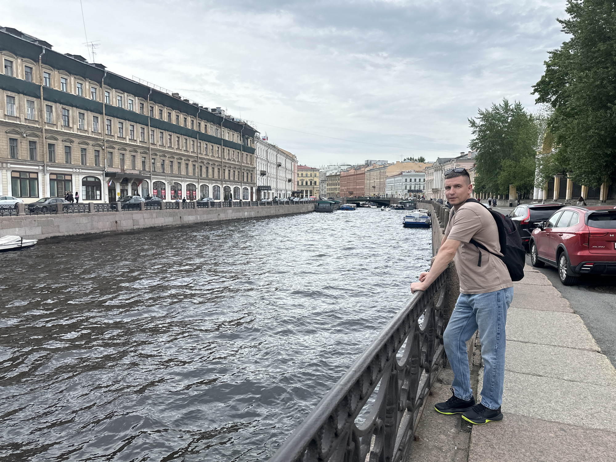 A medium eye-level outdoor shot depicts a man standing on a sidewalk next to a canal. The location is Saint Petersburg, Russia, as suggested by the architecture and waterway scene.
The man, facing slightly toward the camera and with his head turned toward the viewer, leans against a black metal railing. He wears a tan T-shirt, blue jeans, and black sneakers with green accents. A black backpack hangs on his back.
To his right, on the sidewalk, are several parked cars, including a red SUV. To his left, beyond the railing, is a wide canal with gentle ripples on the surface. Boats are sparsely distributed on the water.
On the opposite side of the canal is a row of historic buildings with beige facades and multiple windows. Cars are parked along the street in front of the buildings. In the distance, the buildings become lighter in color, transitioning to whites and yellows.
The sky is overcast with a gray cloud cover. Green trees are visible on the right side of the frame, adding a touch of natural color. The overall lighting is subdued, reflecting the cloudy weather.