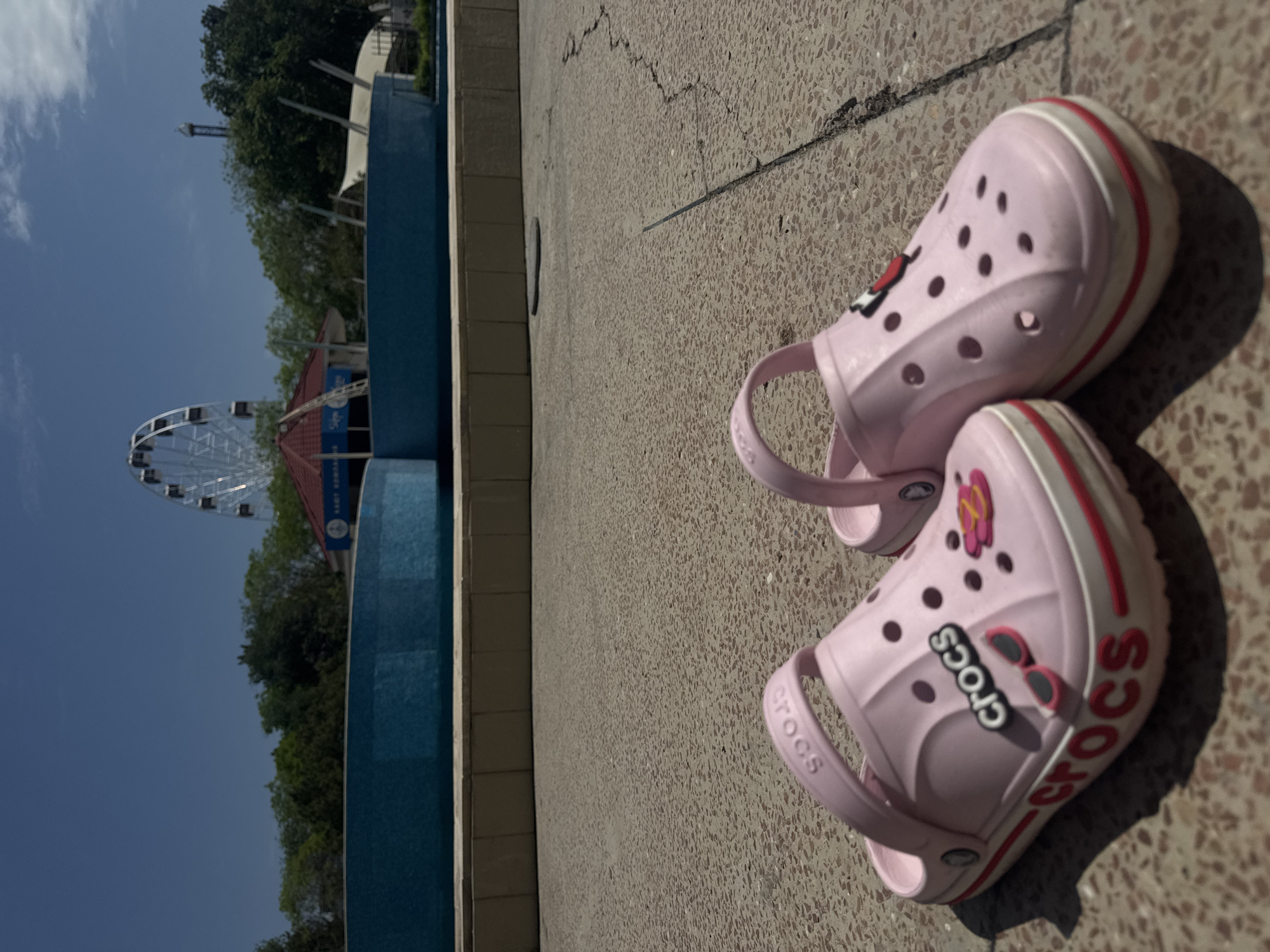 The image shows a pair of pink Crocs sandals on a textured concrete surface. In the background, a Ferris wheel is visible in the distance, located in Yevpatoriya, Ukraine. A blue structure, possibly a pool or wall, is also present in the background. The sky is blue with some clouds.