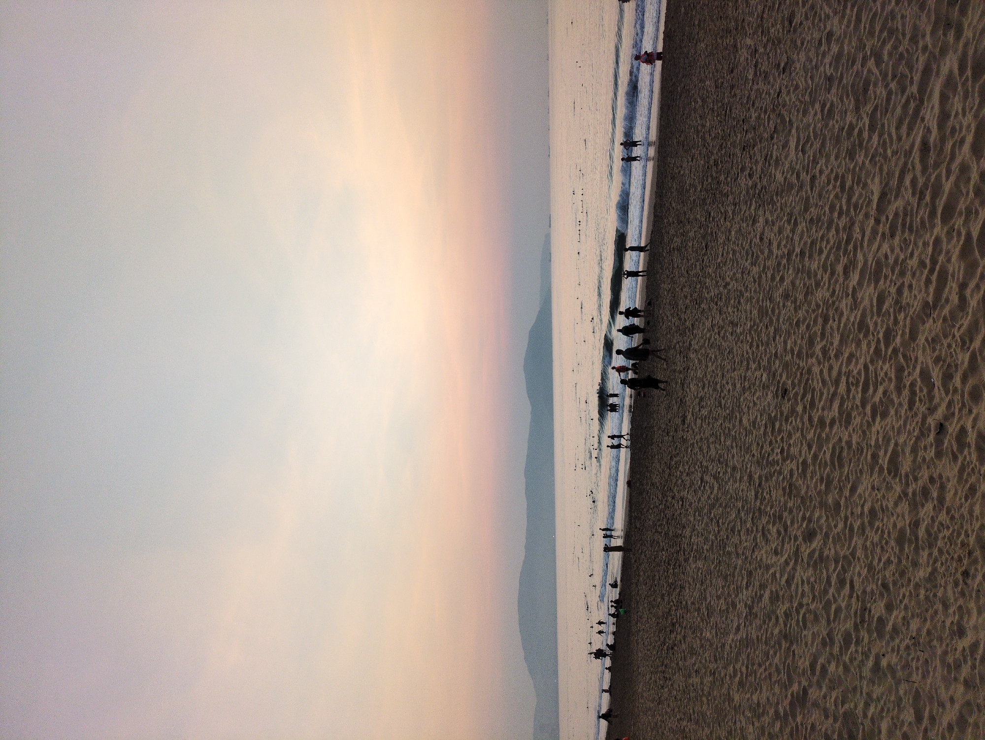 The image depicts a beach scene at sunset. The sky transitions from a light grey to a peach-colored hue. A mountain range is visible in the distance, positioned between the sky and the sea. The sea appears calm with small waves lapping the shoreline. Numerous people are scattered along the beach, both in the water and on the sand. The beach sand is dark and textured. The location is Unknown, Unknown.