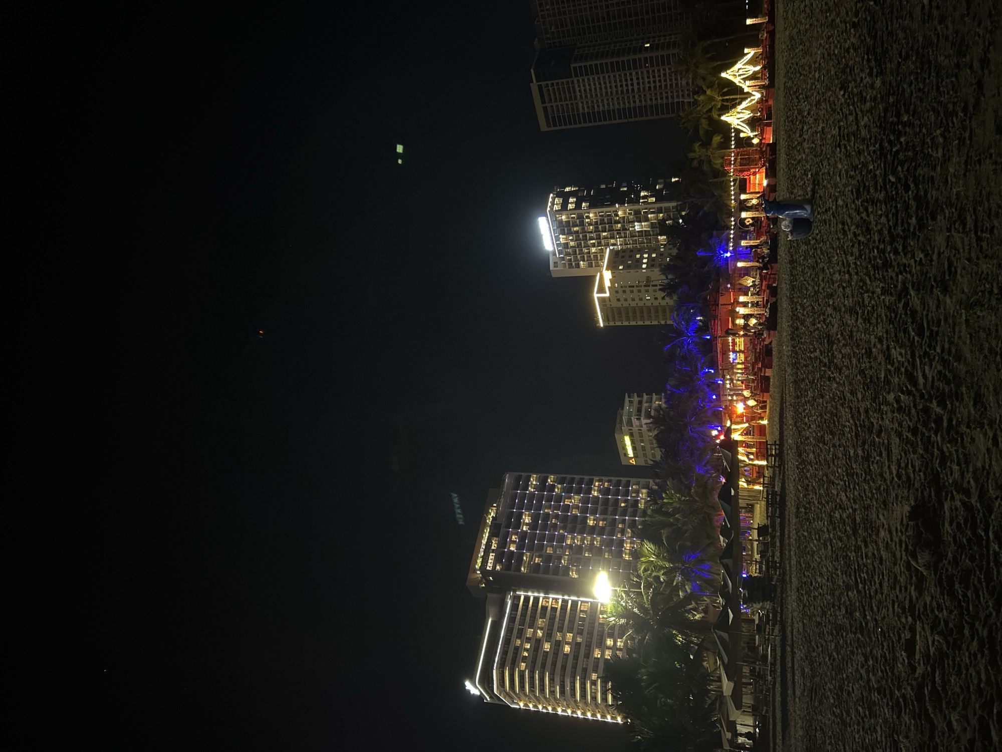 This image captures a nighttime view of a beach in Đà Nẵng, Vietnam, likely near a resort or hotel area. In the background, several brightly lit high-rise buildings stand tall against the dark sky. These buildings feature illuminated windows and decorative lighting along their facades. In the foreground, the sandy beach is visible, with beach chairs and umbrellas arranged in rows. Several small structures or stalls, decorated with festive string lights in red and yellow, are set up on the sand. Palm trees are interspersed throughout the scene, their fronds dark against the illuminated buildings. Two figures are visible on the beach, appearing to be casually dressed and standing near a trash bin. The overall atmosphere suggests a lively evening at a tourist destination.