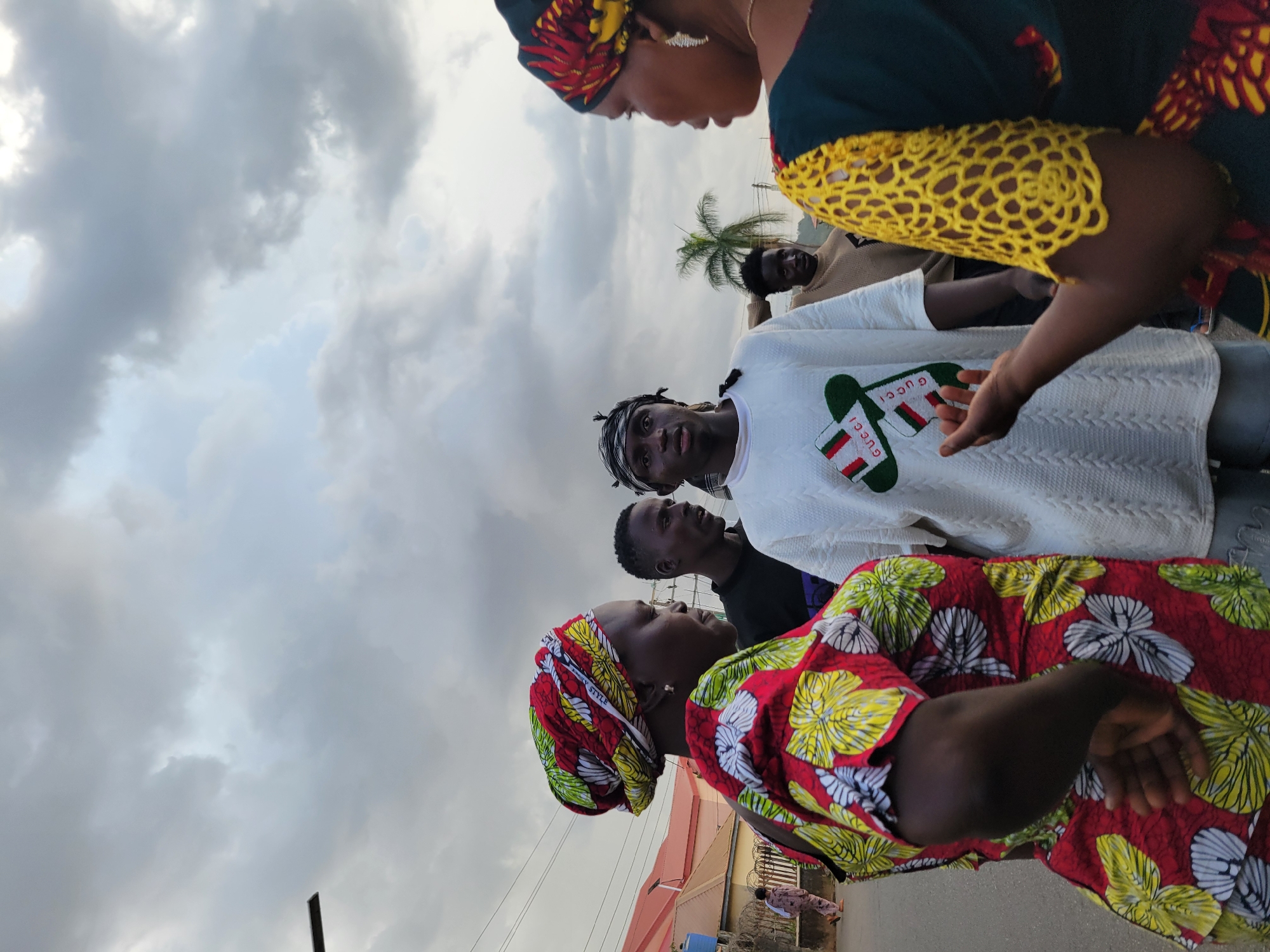In this media file, a group of people are gathered outdoors. The primary subjects are a woman in the foreground wearing a vibrantly patterned headscarf and dress, and a young man in the middle ground wearing a white long-sleeved shirt with "GUCCI" branding visible on it. Behind them, other individuals are also present, though less clearly defined. A palm tree is visible in the background, suggesting a tropical or subtropical climate. The sky is overcast with dramatic clouds, indicating a potentially stormy or humid weather condition. The overall scene appears to be a casual outdoor gathering.
The individuals seem to be interacting, with the woman in the foreground looking upwards and to the side, possibly engaged in conversation. The young man in the white shirt is looking forward with a neutral expression. The time of day is unclear due to the overcast sky, but the lighting suggests it could be daytime.
The setting appears to be urban or semi-urban, with some hints of buildings and possibly power lines in the background. The presence of multiple people in close proximity suggests a social interaction or a communal event. The clothing worn by the people, particularly the bright colors and patterns of the woman's attire, is a notable detail. The visible text "GUCCI" on the man's shirt is also a specific detail that identifies a brand. The overall mood conveyed by the image is one of everyday life and social connection in what appears to be a Nigerian context, given the provided location context of Jos, Nigeria.