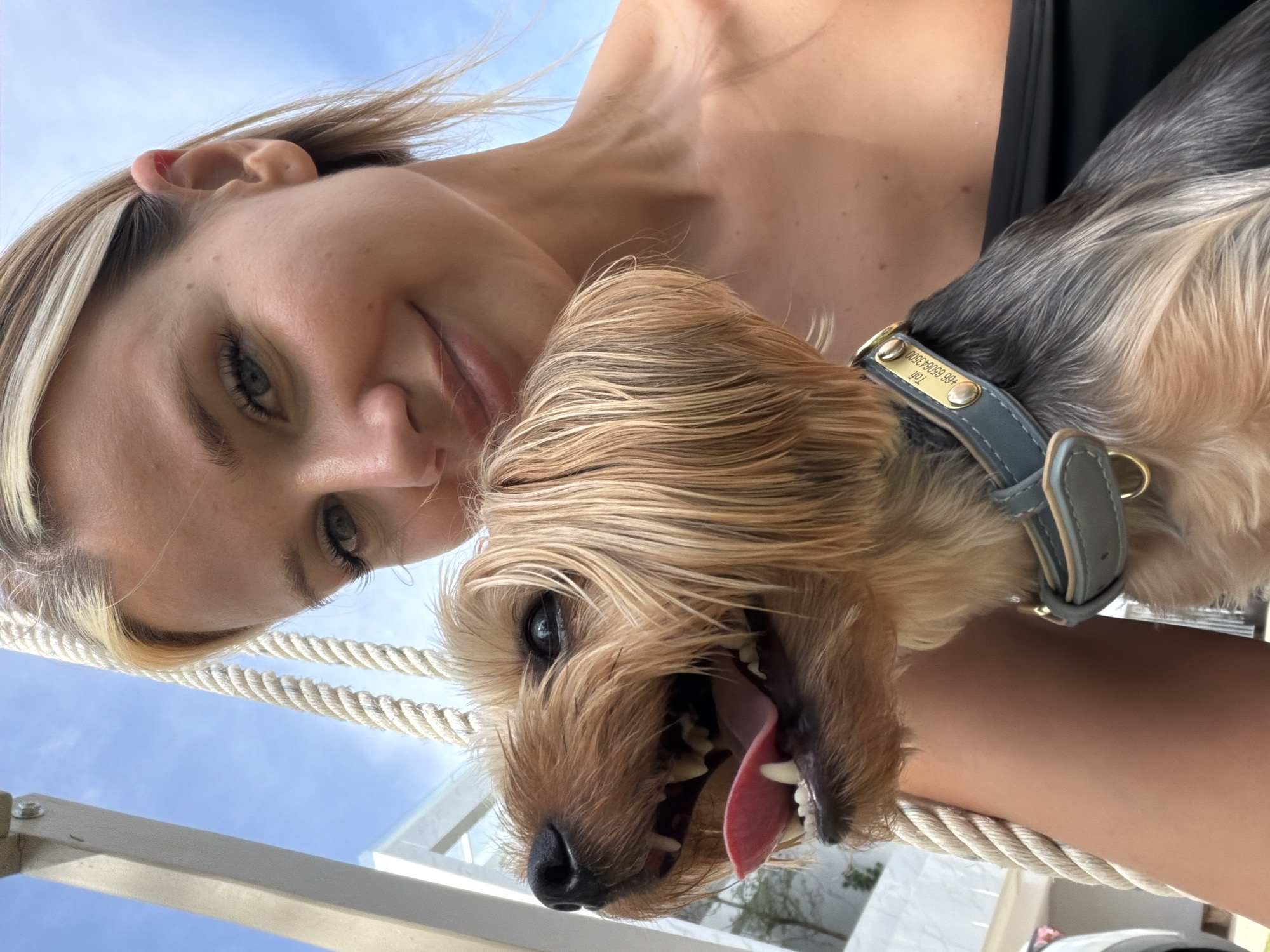 A blonde woman is pictured with her Yorkshire Terrier dog. The dog is in the foreground, with its mouth open and tongue out. The woman is smiling and has her arm around the dog. The background is a blue sky with some clouds, and what appears to be a white building. The setting is outdoors, likely a sunny day. The woman is wearing a black strapless top. The dog is wearing a grey collar with a gold tag. The tag has text on it, including a name and a phone number. The scene appears to be a selfie taken by the woman. The building in the background might be part of a resort or private residence, possibly in an area like Thep Krasatti, Thailand, given the warm climate suggested by the lighting and attire.