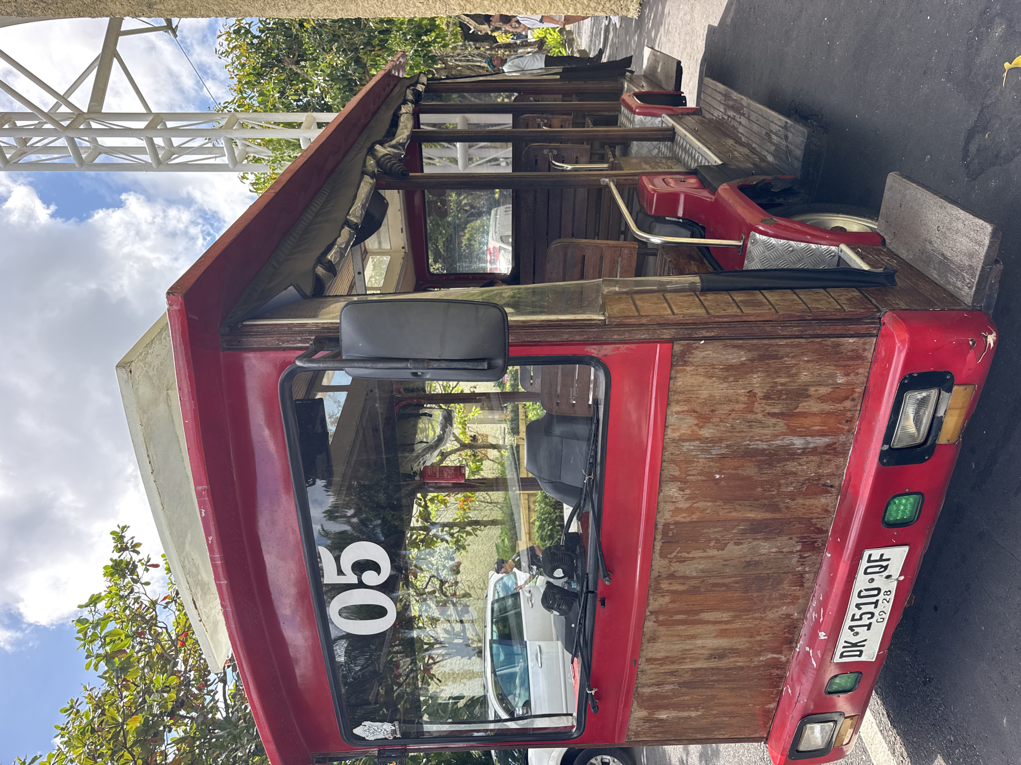 The image shows a red, wooden, open-air vehicle, possibly a modified truck or bus, parked on a street in what appears to be Kuta Selatan, Indonesia based on the vehicle registration plate "DK 1510 QF". The vehicle has a white number "05" displayed on its windshield. It features wooden planks for its body and passenger seating, suggesting a rustic or custom design. The vehicle is parked alongside a paved road next to a curb. In the background, there is foliage, part of a building with white structural beams, and reflections of other vehicles and people in the windshield. The sky is visible with scattered clouds. The image seems to be taken at eye level.
