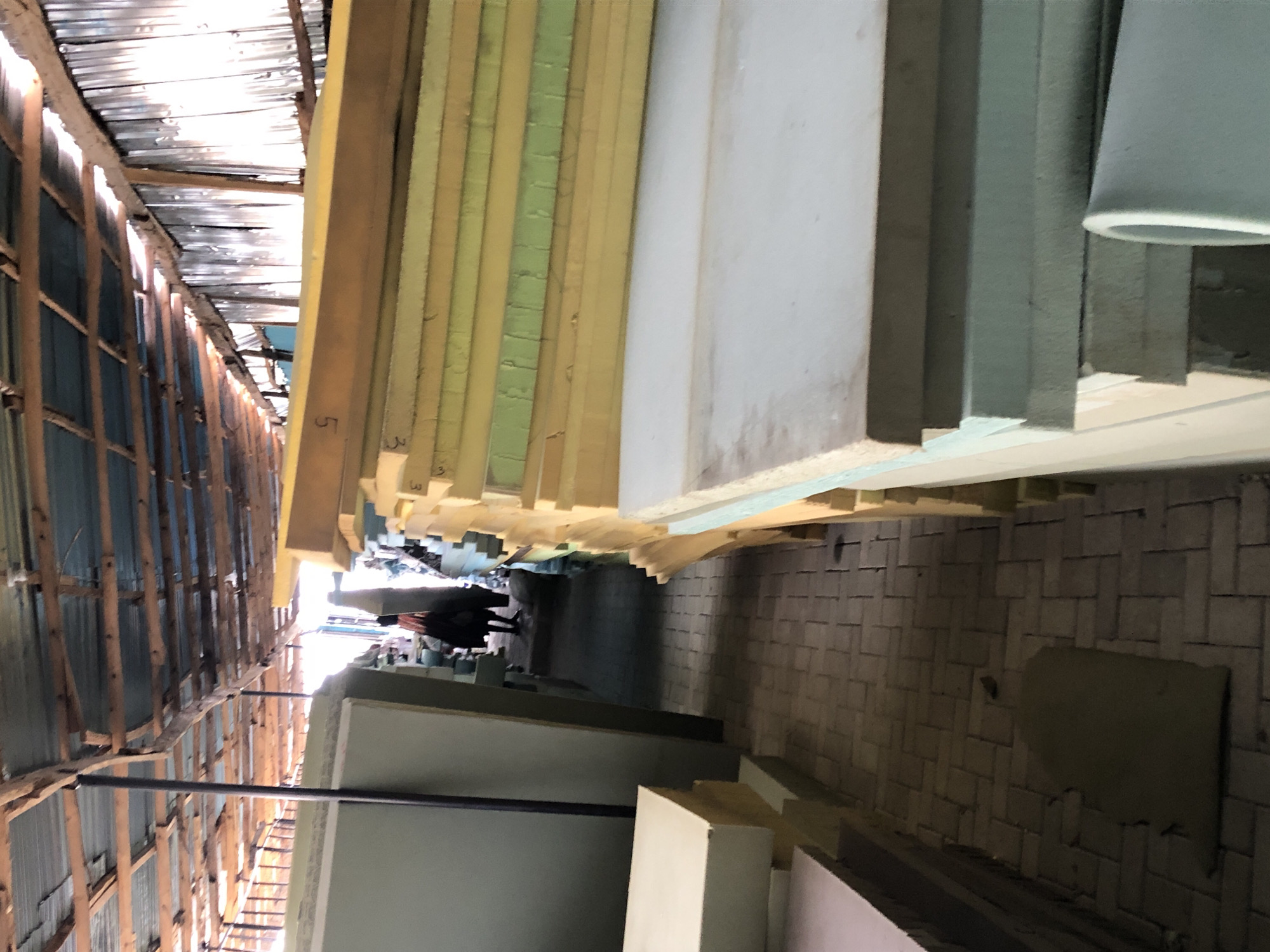The image depicts an indoor space in Ado, Nigeria, potentially a warehouse or storage facility. Stacks of rectangular materials, likely foam or insulation boards of varying thicknesses and colors (yellow, green, white, gray), are piled high on either side of a narrow walkway. The building's interior has a corrugated metal roof supported by wooden beams. The floor is brick. At the far end of the walkway, two figures, possibly people, are walking away from the camera. A black metal bar stretches across the lower left of the frame, likely a support structure. The overall lighting is uneven, with bright areas near the roof and darker areas closer to the floor.