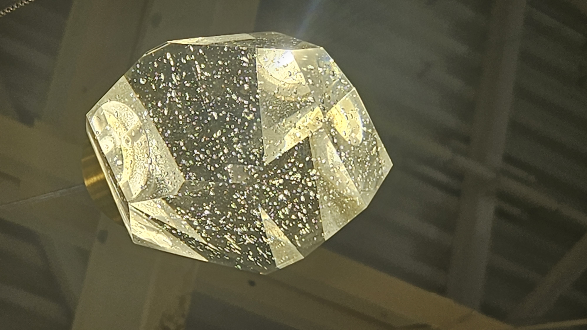 The image shows a close-up of a faceted, clear crystal object, possibly a pendant light or a decorative element, suspended in what appears to be an indoor setting. The object is geometrically shaped, with multiple flat surfaces that catch and reflect light, creating a shimmering effect. Within the crystal, numerous small, sparkling inclusions are visible, contributing to its dazzling appearance. The object is attached to a gold-colored fitting or base, from which it hangs, likely by a wire or cord that is partially visible.
The background is dark and out of focus, suggesting a dimly lit environment. There are indistinct structural elements visible, such as beams or a ceiling structure, possibly made of metal or wood, with a ribbed or corrugated texture. These elements, combined with the lighting, create a somewhat industrial or architectural backdrop. The overall impression is one of modern or contemporary design. There is no visible text in the image. The lighting conditions suggest it might be evening or an interior space with artificial illumination. No people or animals are present in the frame.