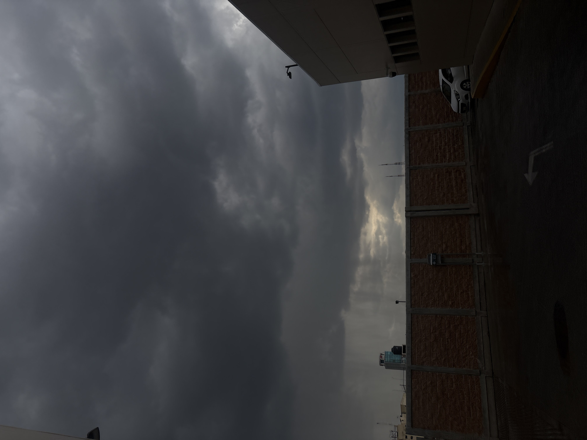 The image shows a cloudy sky above what appears to be a parking lot and the side of a building in Puebla, Mexico. The sky is predominantly dark gray with a lighter, brighter area visible toward the center. A tall brick building extends along the right side of the image, adjacent to an asphalt surface with a directional arrow painted on it. A white car is parked along the building. In the background, distant buildings and antennas are visible against the sky. Surveillance cameras are attached to the building's exterior.