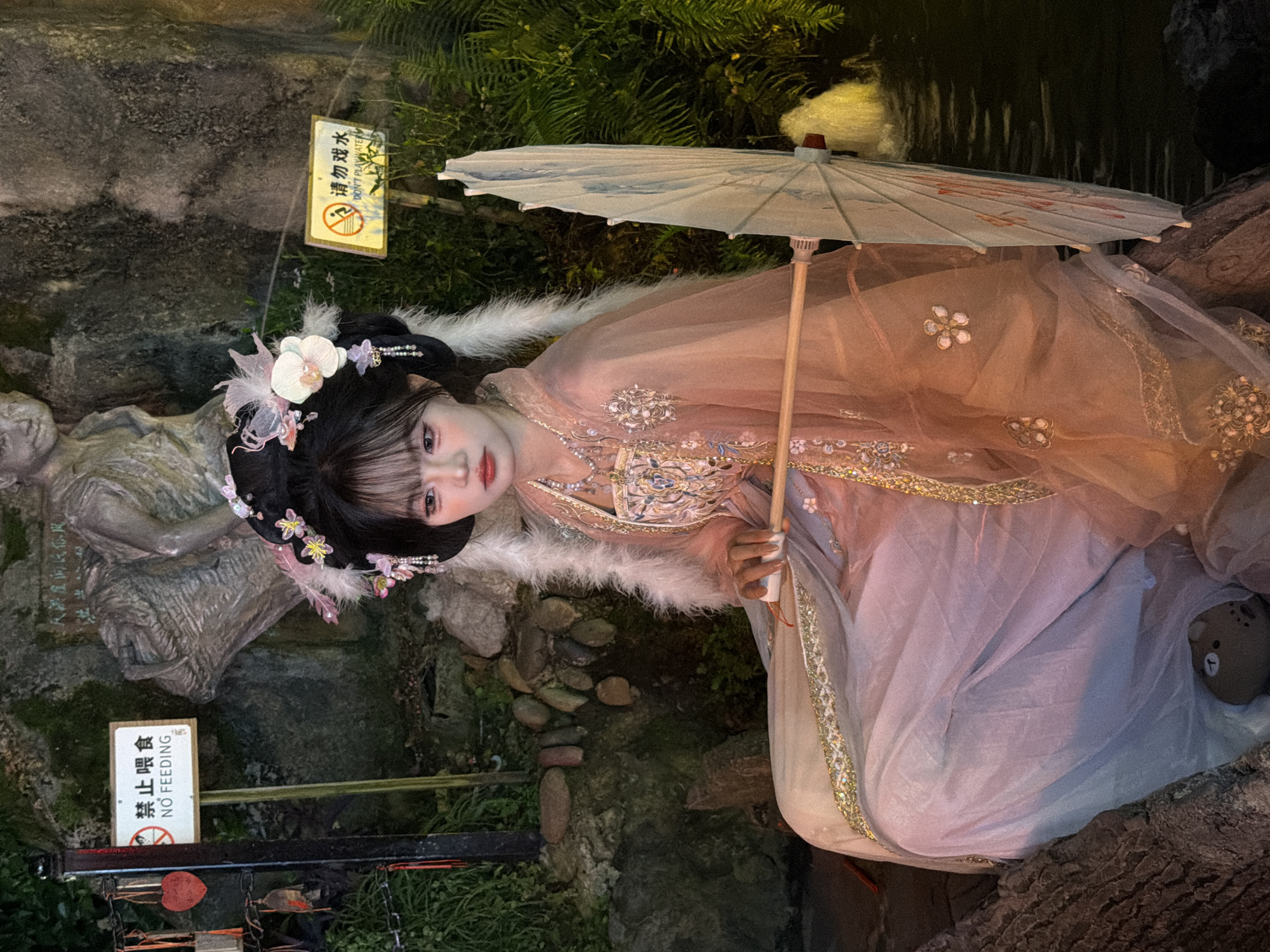A young woman in traditional Chinese attire, featuring a pink and pastel dress with intricate embroidery and a feathered collar, is seated outdoors. Her hair is styled elaborately with flowers and ornaments. She holds a closed, light-colored umbrella with a wooden handle. The setting appears to be a park or garden, with a stone wall, lush greenery, and a body of water visible in the background. Two signs are present: one with Chinese characters and "NO FEEDING" in English, and another with Chinese characters and "DON'T PLAY WATER". The lighting suggests it is evening or nighttime.