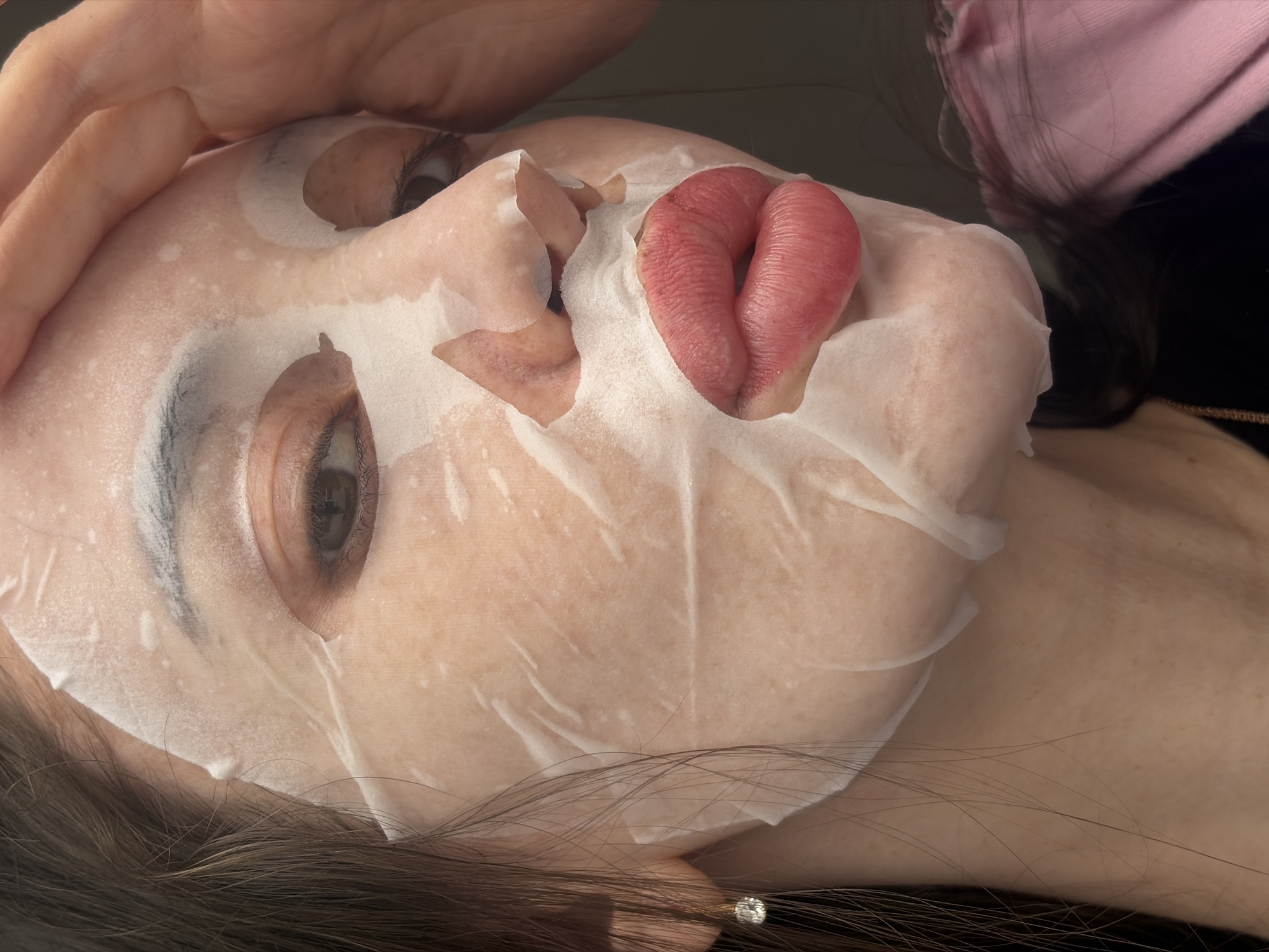 A close-up, tilted, indoor shot features a person wearing a facial mask. The mask is torn around the mouth area, revealing a protruding tongue. One hand is visible, resting on the person's forehead. The background is blurred, but suggests the presence of another person and clothing. The location is not discernable from the image alone.