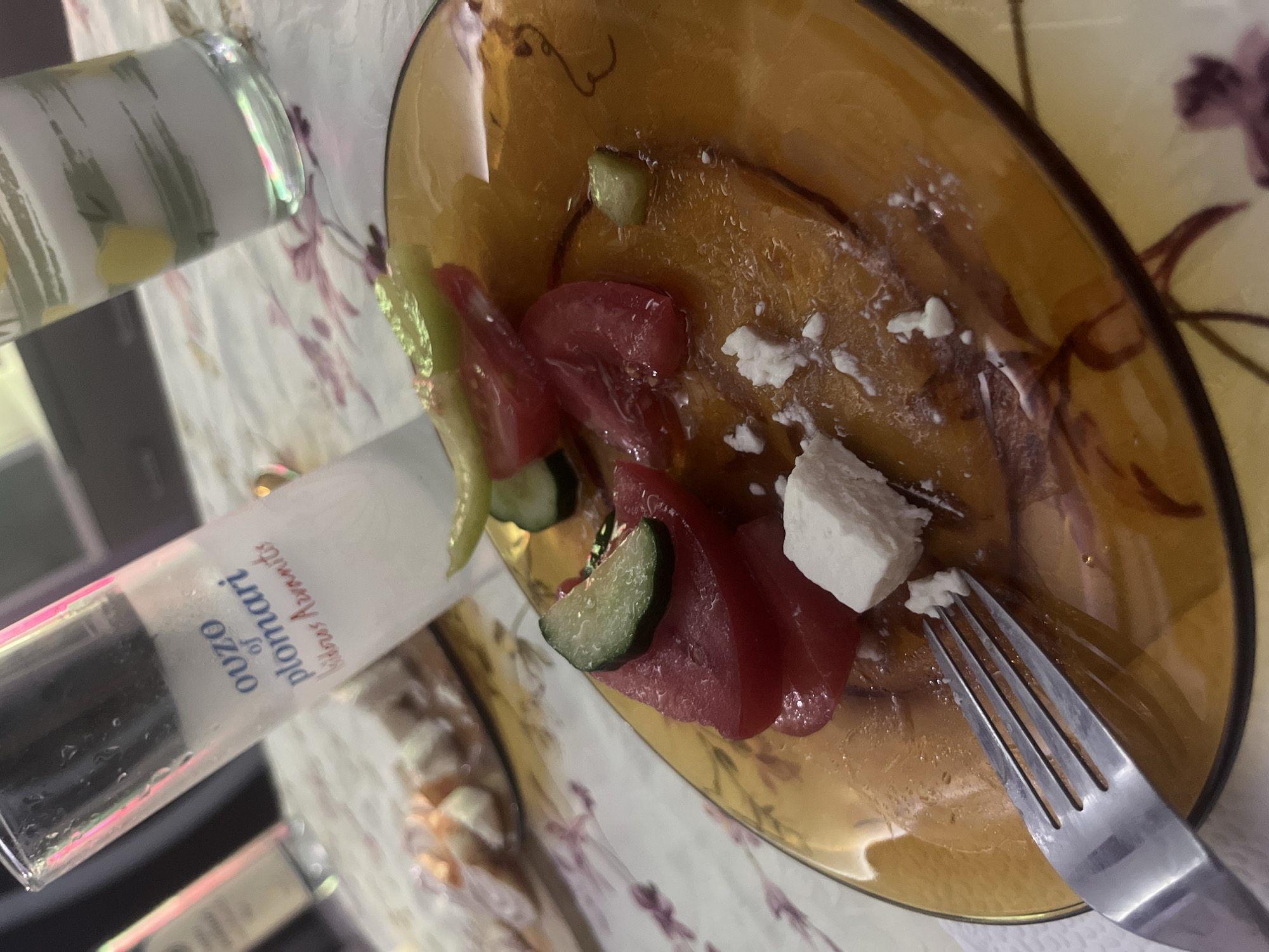The image shows a close-up of a plate of food and a glass of ouzo. The plate contains sliced tomatoes, cucumber, and feta cheese, with a fork resting on the side. The glass is partially filled with a cloudy white liquid, and a label reads "ouzo of plomari". In the background, another glass with a yellow and green liquid is visible, along with a bottle. The scene appears to be set on a table with a floral tablecloth. The image was taken in Burgas, Bulgaria.