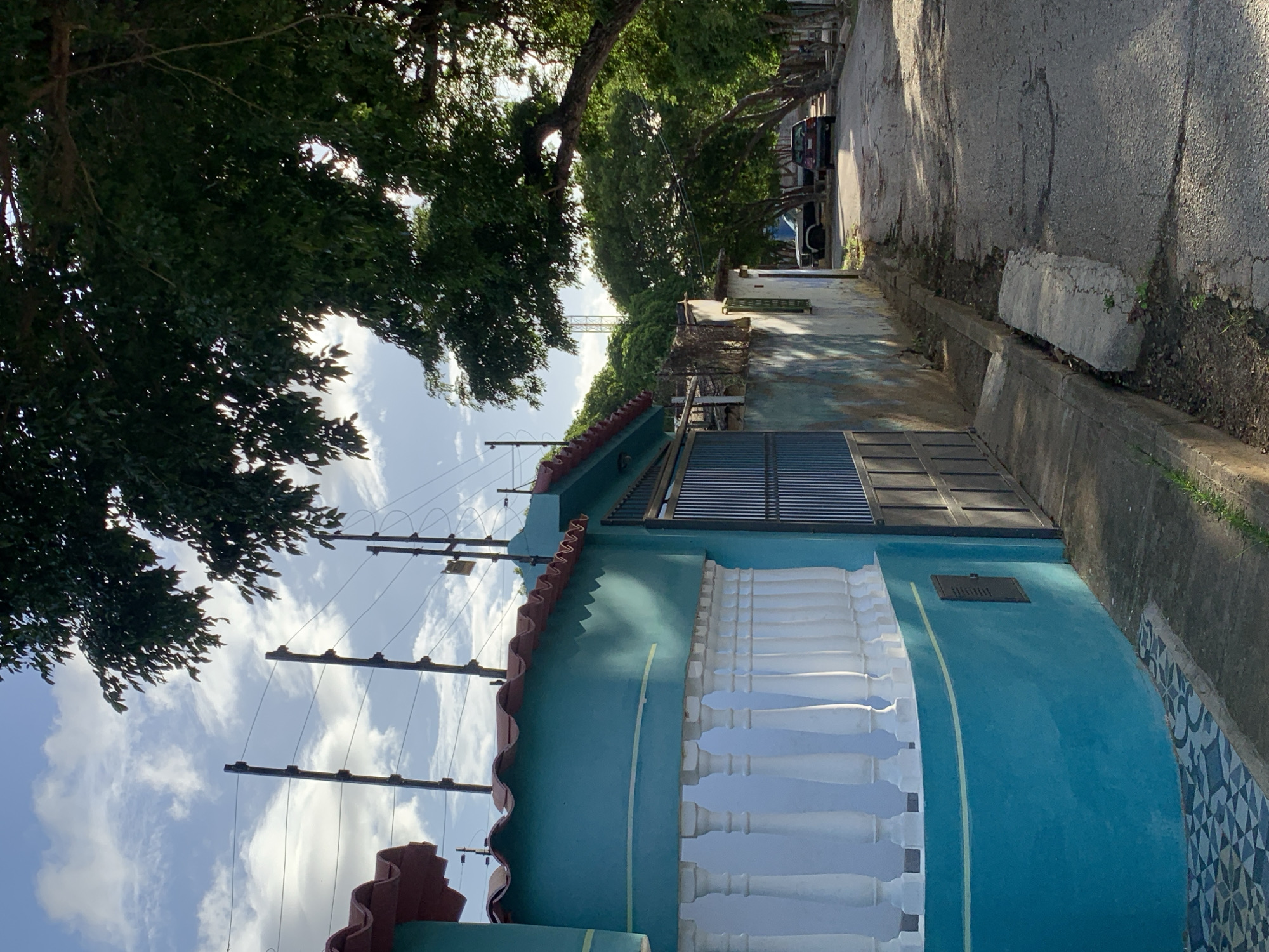 The image shows a street scene in Santa Ana, Venezuela. In the foreground, a bright turquoise building with a red tiled roof features a white balustrade along its curved facade. To the right, a metal gate leads into the property. The street is paved and runs between buildings and mature trees that line the right side of the road, casting shadows. Several cars are parked along the street, and utility poles with electrical wires are visible in the background against a partly cloudy sky. The scene appears to be captured during daylight.