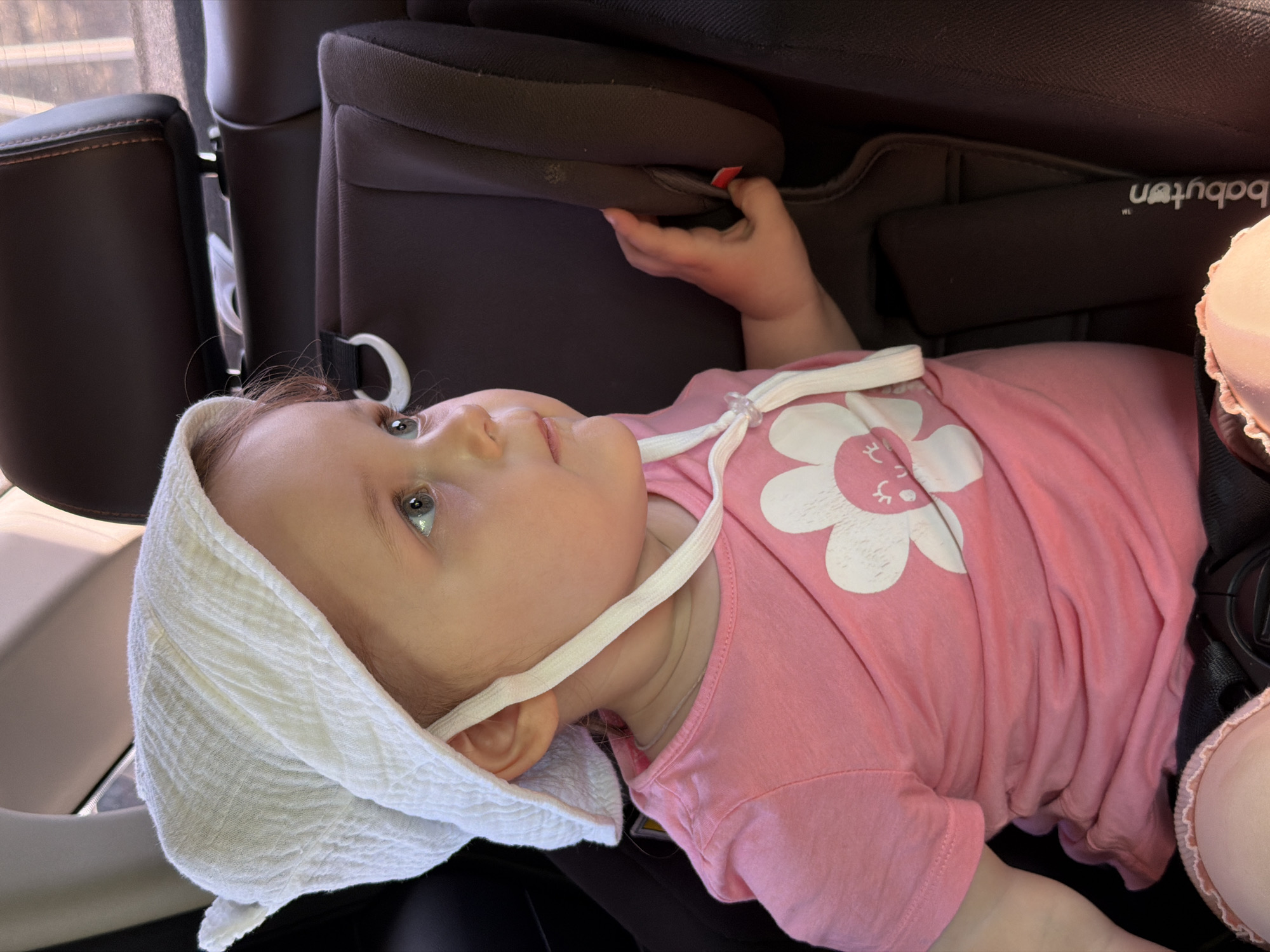 A baby wearing a pink shirt with a white flower design and a white bonnet is lying in a black car seat. The car seat is situated in the backseat of a vehicle. The location and city of Chaikovsky, Russia, are not determinable from the image.