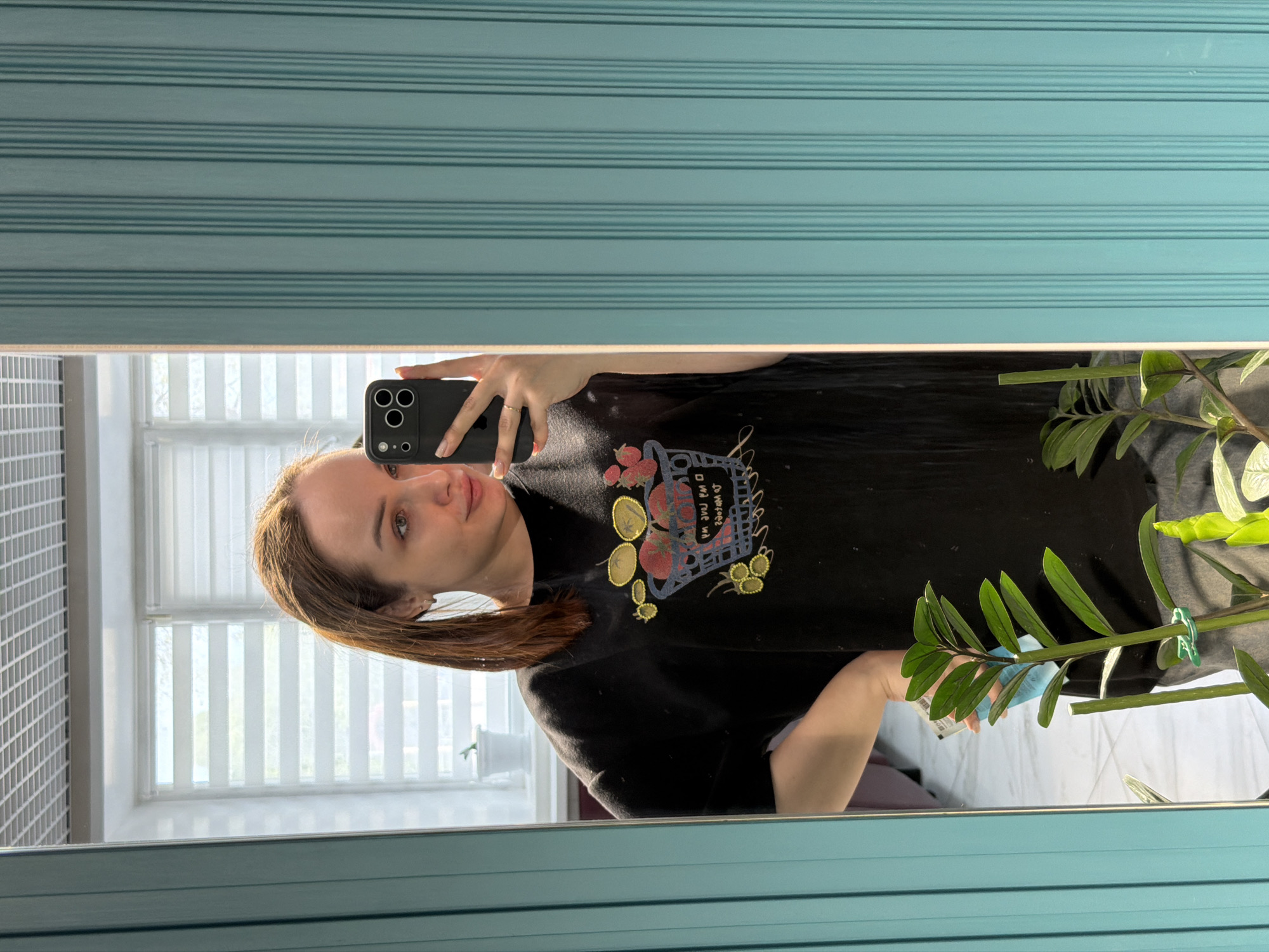 A woman is taking a mirror selfie in what appears to be a room with a window and a houseplant. The window has white blinds, and there is a grid-like pattern on the wall to the left of the window. The mirror is framed with a light blue border with horizontal lines. The woman is holding a black smartphone and wearing a black t-shirt with a graphic design. She has brown hair and is looking at the phone. A green houseplant is visible in the lower right corner of the mirror. The location is Chaikovsky, Russia.