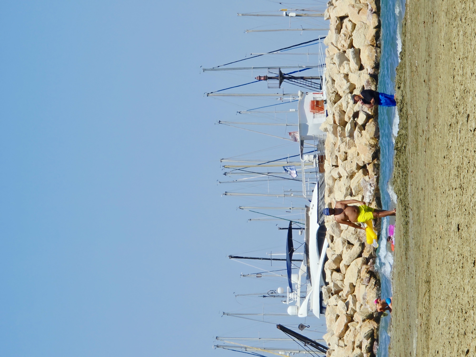 The image depicts a sunny day at a beach adjacent to a marina in Larnaca, Cyprus. In the foreground, the sandy beach is occupied by several individuals. A man in yellow shorts walks towards the right, carrying a yellow inflatable toy. To his left, a child in blue shorts is playing in the shallow water. Further to the left and slightly in the background, another child is visible with a pink inflatable ring. To the right of the man in yellow shorts, another person stands in the water, wearing a black t-shirt and blue shorts.
Behind the beachgoers, a rocky breakwater forms a barrier between the sea and the marina. Beyond the breakwater, a marina is filled with numerous sailboats and yachts moored side-by-side. The masts of these vessels create a dense, vertical pattern against the clear blue sky. A white yacht with a dark blue canopy is prominently featured in the middle ground, partially obscured by the breakwater. A red and white navigation light is visible atop a structure within the marina. The overall scene suggests a leisurely day at the coast with recreational activities taking place both on the beach and in the water, while the marina represents a hub for maritime activities.