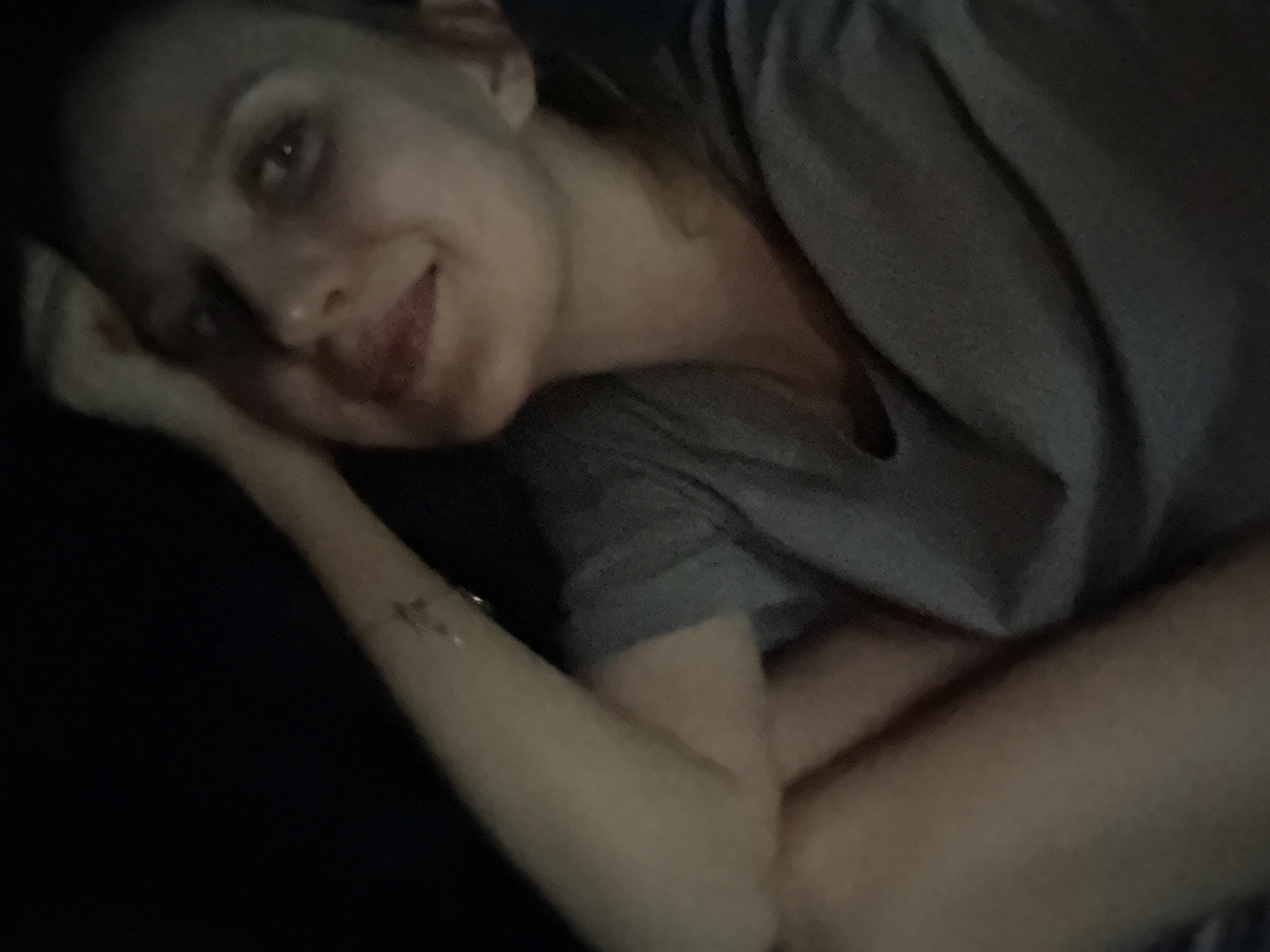 A person with fair skin lies on a dark surface. The person is wearing a gray shirt with a v-neck. The person's head rests on their arm. The location is unknown. There are no identifiable landmarks or details suggesting the city of Izhevsk, Russia.