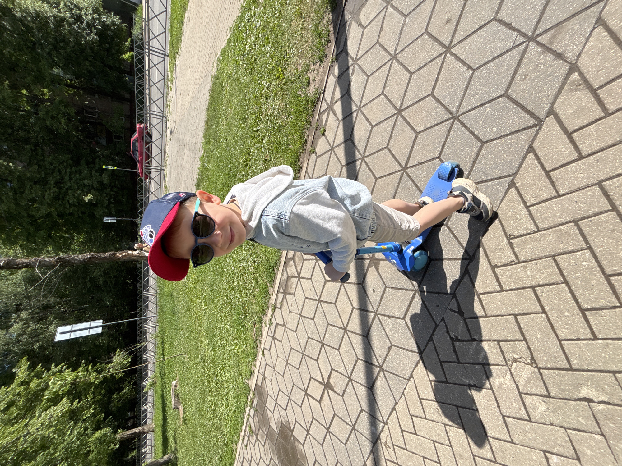 A young boy stands upright on a blue scooter on a sunny day in Rusanovo, Russia. The boy, wearing a red and blue baseball cap, sunglasses, a light-colored hoodie over a t-shirt, and shorts, is positioned centrally on a paved sidewalk. He is looking towards the viewer with a slight smile.
The sidewalk features two distinct patterns: a section closer to the boy has a geometric, optical-illusion cube design, while further down, it transitions to a herringbone brick pattern. Bright sunlight casts long, distinct shadows of the boy and scooter onto the pavement, indicating a clear, sunny sky, likely during midday or early afternoon.
To the boy's left, a strip of vibrant green grass runs alongside the sidewalk, bordered by a metal fence. Beyond the fence, a road is visible where a red car is parked or driving. Lush green trees line the background, with hints of a building structure partially obscured by the foliage. A white rectangular street sign on a pole is also visible in the grassy area. No discernible text is visible on any object or surface in the image.