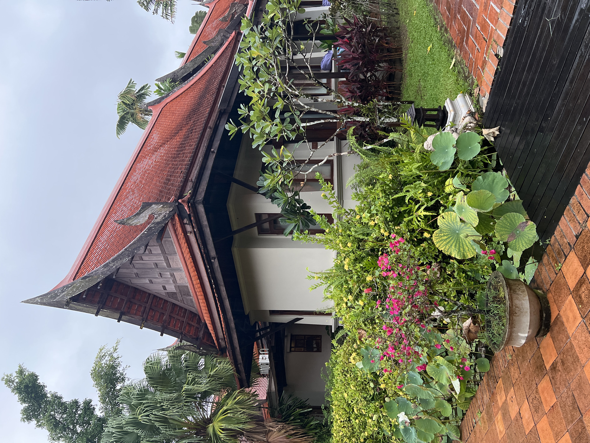 A Thai-style building with a red tiled, multi-tiered roof sits behind a lush garden overflowing with green foliage, pink and yellow flowers, and large lily pads. The building's facade is cream-colored with dark wooden trim around the windows. A brick pathway, damp from recent rain, leads to the building. In the foreground, a dark wooden walkway is also wet. The sky is overcast and the scene appears to be in a resort or hotel setting in Ban Bang La On, Thailand.