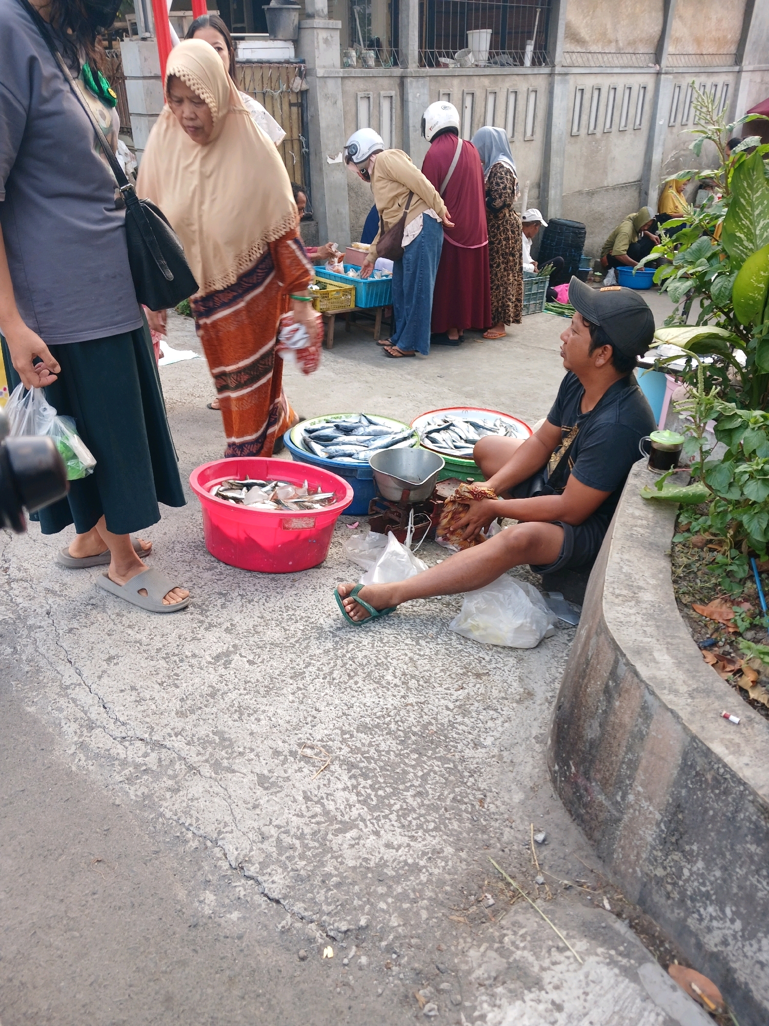 The image depicts an outdoor market scene in Banyuwangi, Indonesia, possibly near a roadside or sidewalk. In the foreground, a man wearing a black baseball cap and a dark t-shirt is sitting on the ground, holding a small bundle of something in his hands. He appears to be a vendor, with several plastic bags of goods scattered around him. To his left, a bright pink bucket overflows with small fish, and next to it, a larger, shallow metal tray also contains fish. Behind him, other vendors are visible, including a woman in an orange and brown patterned dress and a light brown hijab, who is holding a white plastic bag. Other people in the background are either standing or seated, and some are wearing helmets, suggesting transportation by motorcycle. The ground is paved with a textured, grey concrete surface, showing some cracks. The environment is somewhat cluttered, with various market items and debris present. A concrete curb runs along the right side of the image, with some vegetation growing nearby. The overall atmosphere suggests a busy and informal marketplace.