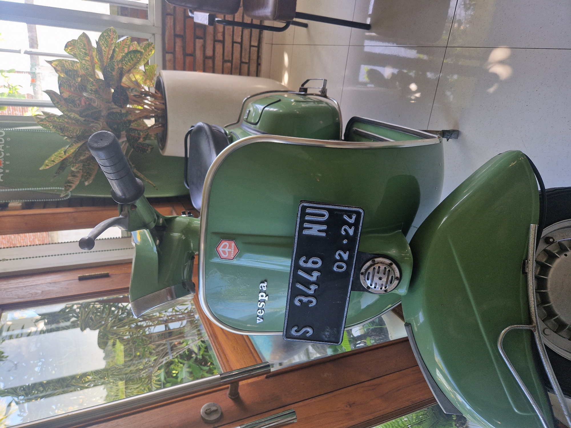 A green Vespa scooter is parked indoors near a window with wooden frames. The license plate number is S 3446 NU, valid until 02-24. A potted plant is behind the scooter. The location is Pekutatan, Indonesia.