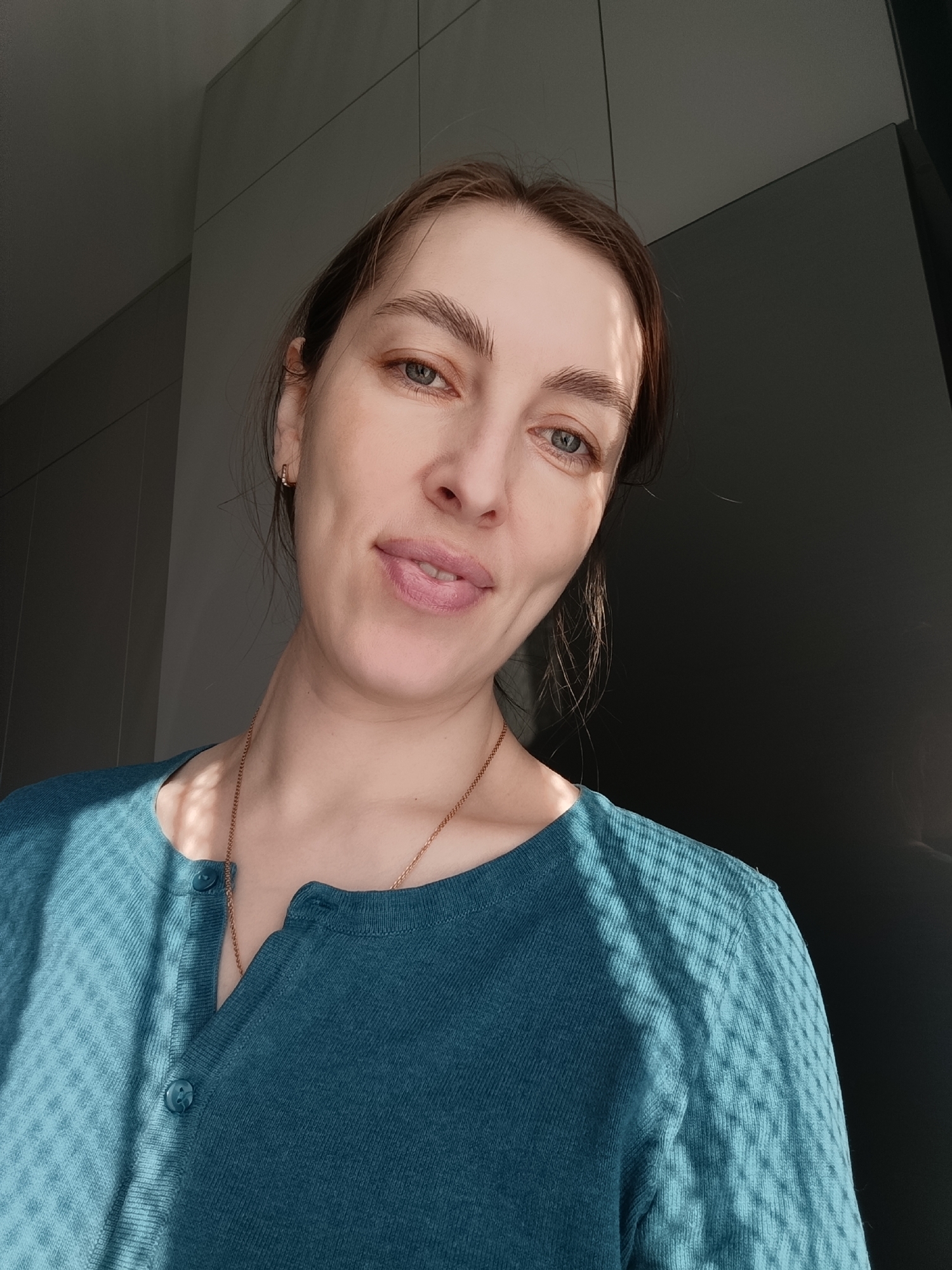 A woman is posing for a selfie indoors. She has brown hair and light blue eyes. She is wearing a teal blue cardigan with buttons and a gold chain necklace. There is light falling across her shirt in a patterned, grid-like fashion. She is positioned against a grey and dark grey background, likely a wall or cabinet. The image was taken in Chaikovsky, Russia.