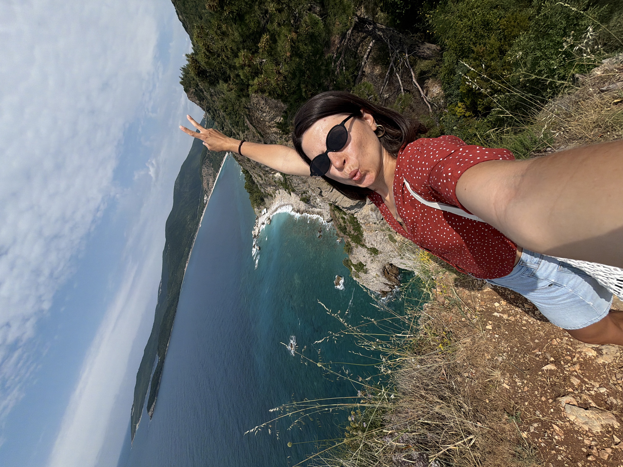 A woman is captured in a selfie taken from a high vantage point overlooking a beautiful coastal landscape in Budva, Montenegro. The image is rotated, suggesting the woman is holding the camera horizontally, perhaps at arm's length.
The subject is a woman with dark hair, wearing sunglasses, small hoop earrings, a red top with white polka dots, and light blue denim shorts. A white woven bag strap is visible across her chest. She has one arm extended towards the left, making a peace sign with her fingers, while her other arm is extended holding the camera. Her facial expression shows a slight pout.
The scene unfolds from a cliff edge, looking down towards a bay with clear turquoise and dark blue waters. Rocky shorelines and small beaches are visible below, with waves gently breaking. In the distance, a long stretch of land with dense green vegetation extends into the sea, forming the other side of the bay. The sky above is bright and mostly clear, with scattered white clouds. The immediate foreground around the woman is covered in dry grass and sparse vegetation, characteristic of a Mediterranean coastal area. The time of day appears to be sunny daylight. No visible text is present in the image.