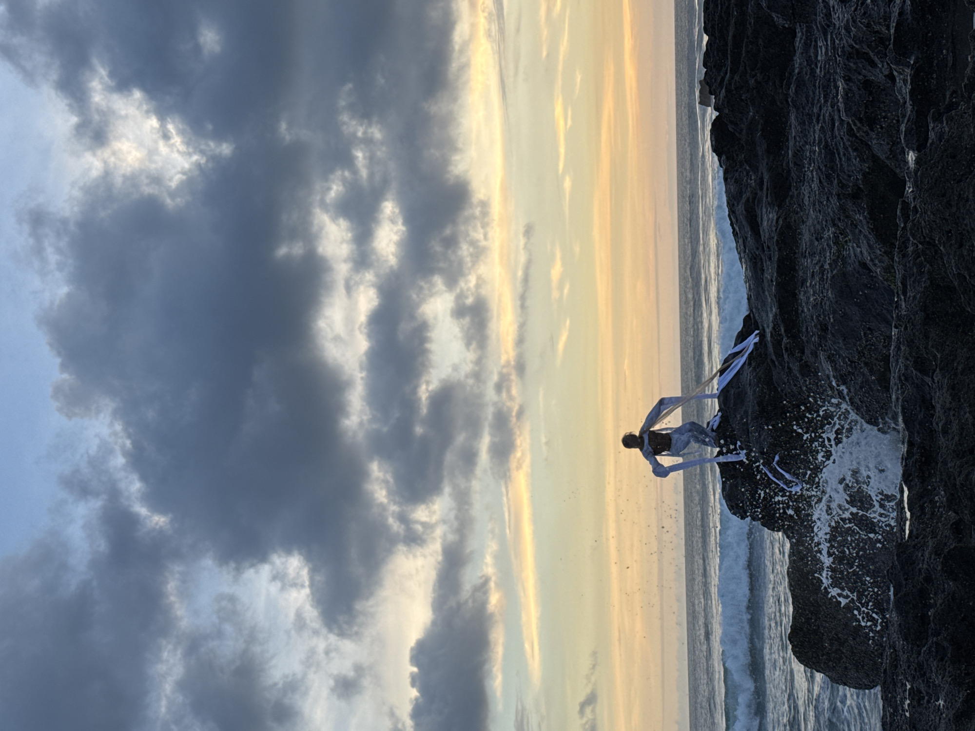 A dramatic coastal scene captured at either sunrise or sunset in North Kuta, Indonesia. The image, which appears to be rotated 90 degrees clockwise, presents a vast and dynamic sky, a rugged coastline, and an ocean with crashing waves.
The sky, occupying the left side of the (mentally corrected) frame, transitions dramatically. On the far left, heavy, dark grey clouds dominate, with hints of lighter sky peeking through. Towards the right, where the sun is setting or rising, the clouds dissipate, revealing brilliant golden and soft yellow light stretching across the horizon.
In the foreground, a massive, dark, and highly textured rock formation occupies the right side of the image. Its jagged and uneven surface suggests volcanic origins. Waves are actively crashing against the base of these rocks, creating white, frothy foam and spray that contrast sharply with the dark rock.
Perched atop these rugged rocks, silhouetted against the warm glow of the sky and the ocean, is a lone figure. This person appears to be wearing or adorned with light-colored, flowing fabric or ribbons that cascade around them, adding a sense of movement and grace. Their pose is dynamic, possibly mid-action like a jump or a dramatic gesture, suggesting an artistic performance, a photoshoot, or a ritual taking place. The exact features of the person are obscured by the silhouette.
The ocean extends horizontally behind the rocks, with a calmer expanse further out and more turbulent water near the shore where waves break. The overall atmosphere is one of grandeur and natural power, enhanced by the striking light conditions. No text is visible in the media.